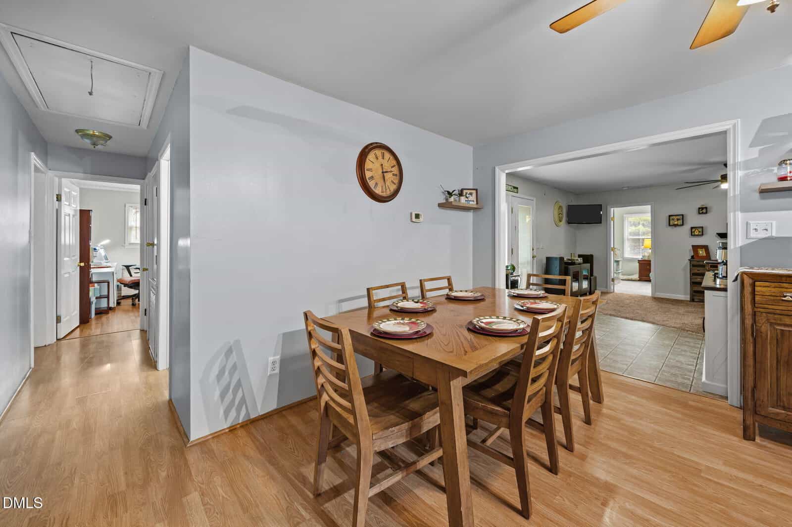 A dining area with a wooden table set for six, wooden chairs, a wall clock, and light wood flooring. The room connects to a hallway, living area, and kitchen space, all with neutral-colored walls and decor.