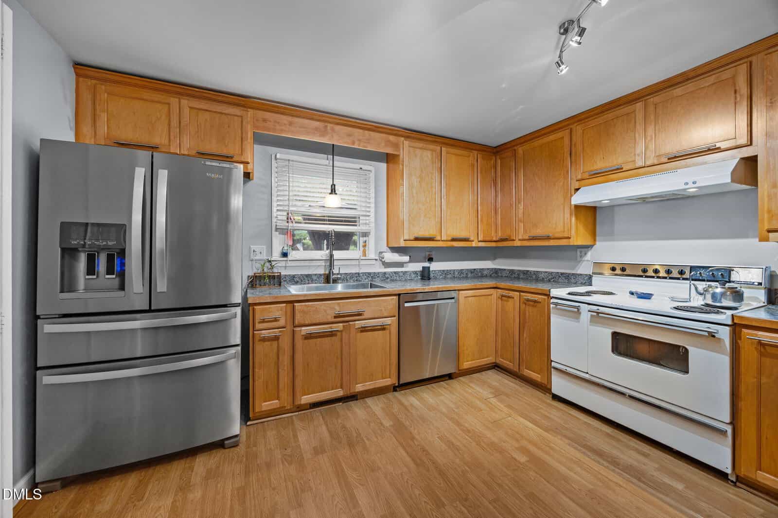 Modern kitchen with stainless steel refrigerator, dishwasher, and sink under a window. Wooden cabinets and drawers line the walls. There is a white stove and oven, and wood flooring throughout the space.