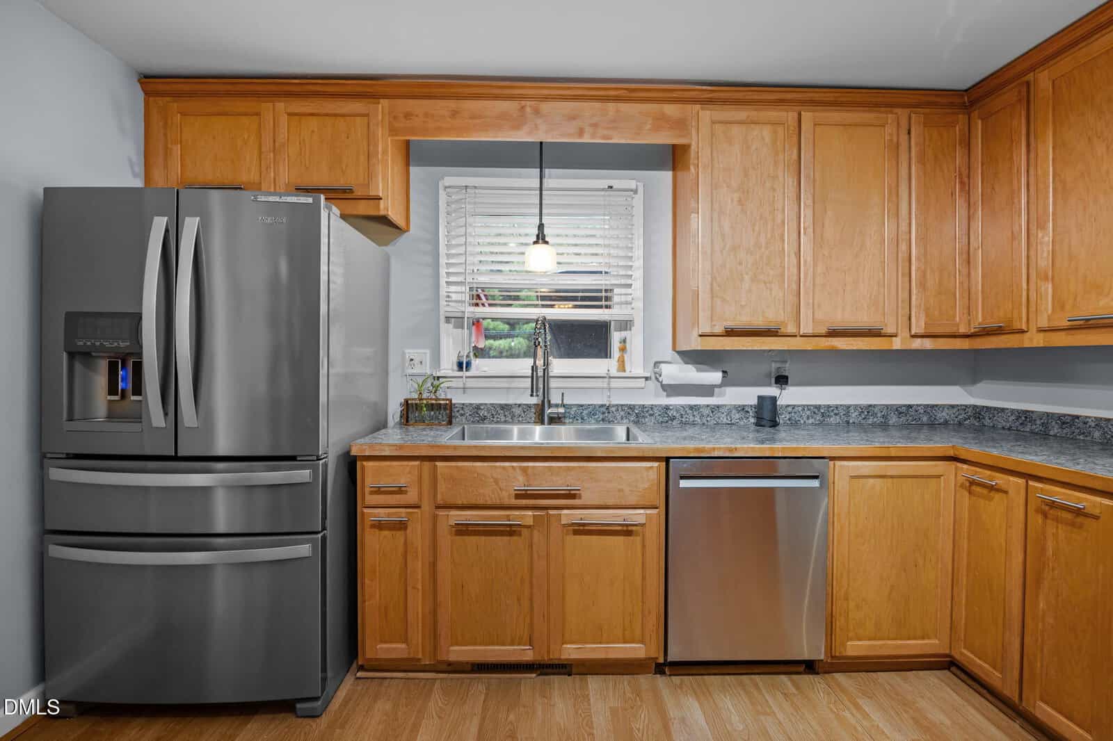 A kitchen with wood cabinets, stainless steel refrigerator and dishwasher, gray countertops, a sink under a window with blinds, a hanging light fixture, and light wood flooring.
