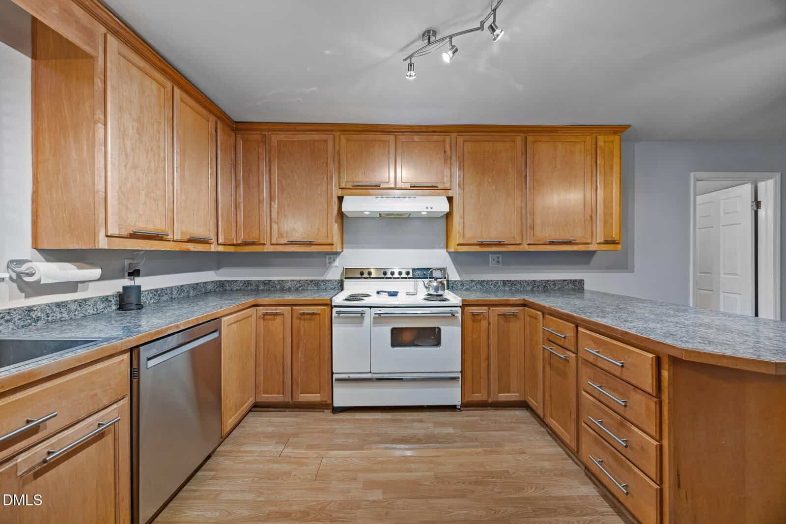 Modern kitchen with light wood cabinets, gray countertops, white stove and oven, stainless steel dishwasher, under-cabinet lighting, and wood floor. Overhead track lighting illuminates the space. Doorway visible on the right.