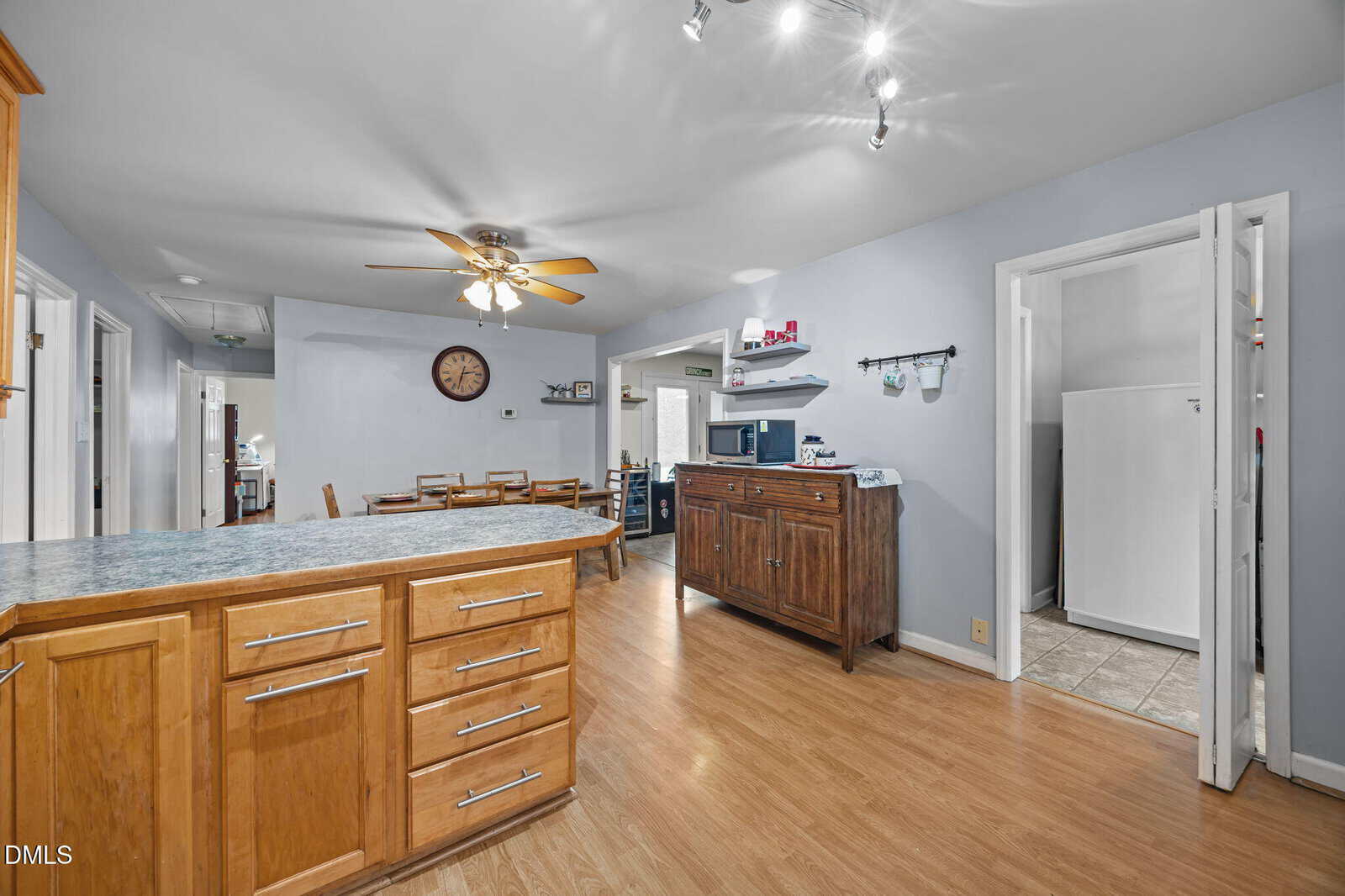 Spacious kitchen with wood cabinets, laminate countertops, and light wood flooring. Dining area with table and chairs, ceiling fan, clock on the wall, and a doorway leading to a utility room with a white appliance.