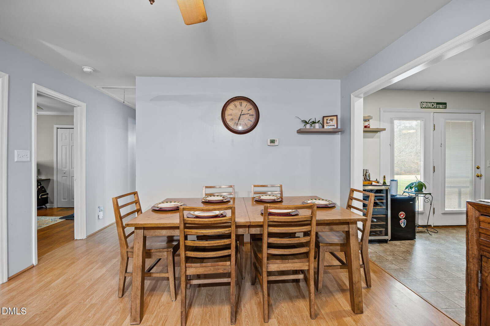 A wooden dining table with six chairs, set with plates and napkins, sits against a light gray wall with a clock above it. The room features wood flooring and an adjacent tiled area with doors and windows.