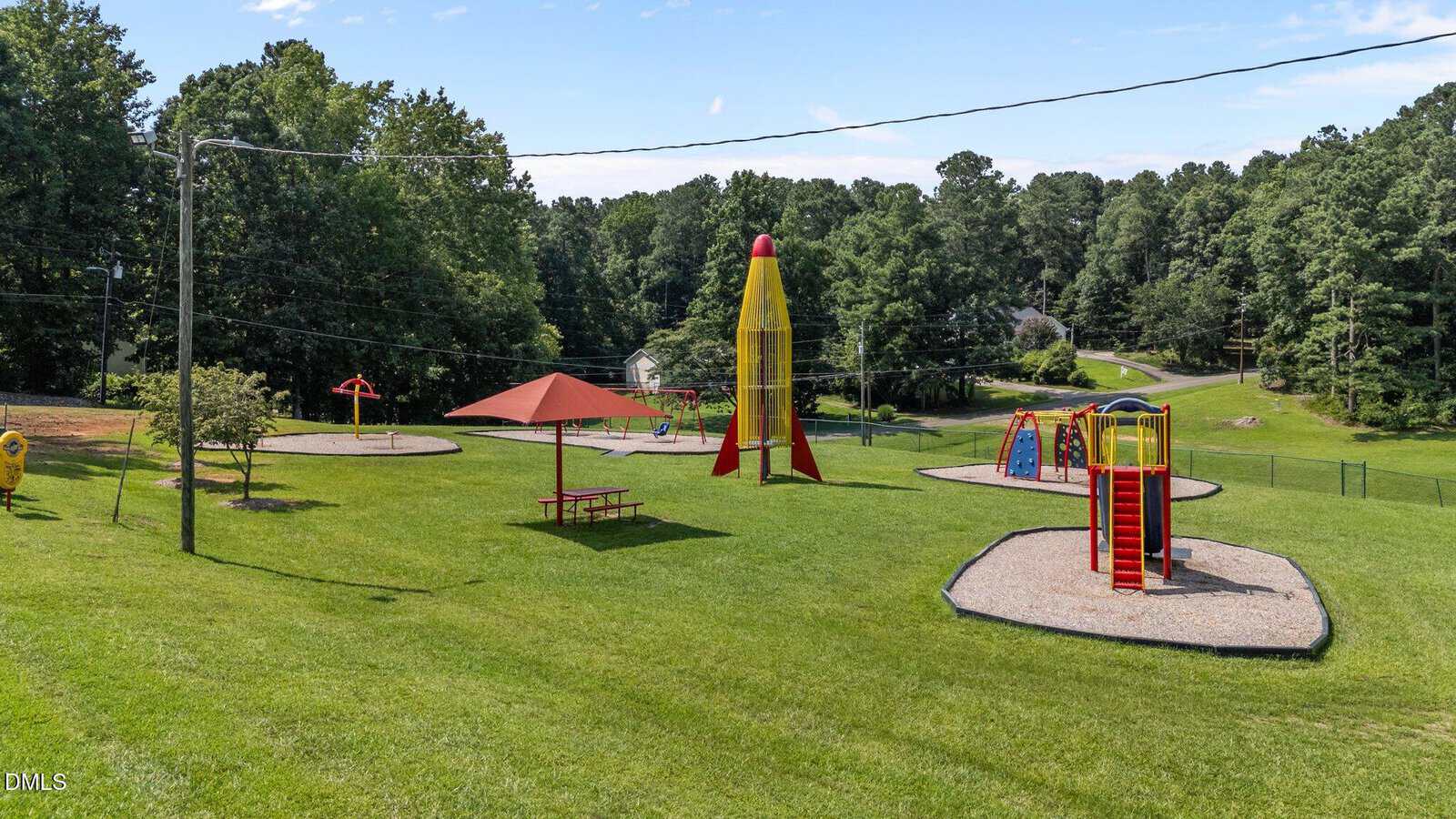 A grassy playground with multiple play structures, including a tall yellow rocket, slides, climbing equipment, a red picnic table under a canopy, and surrounding trees in the background. Power lines run across the scene.