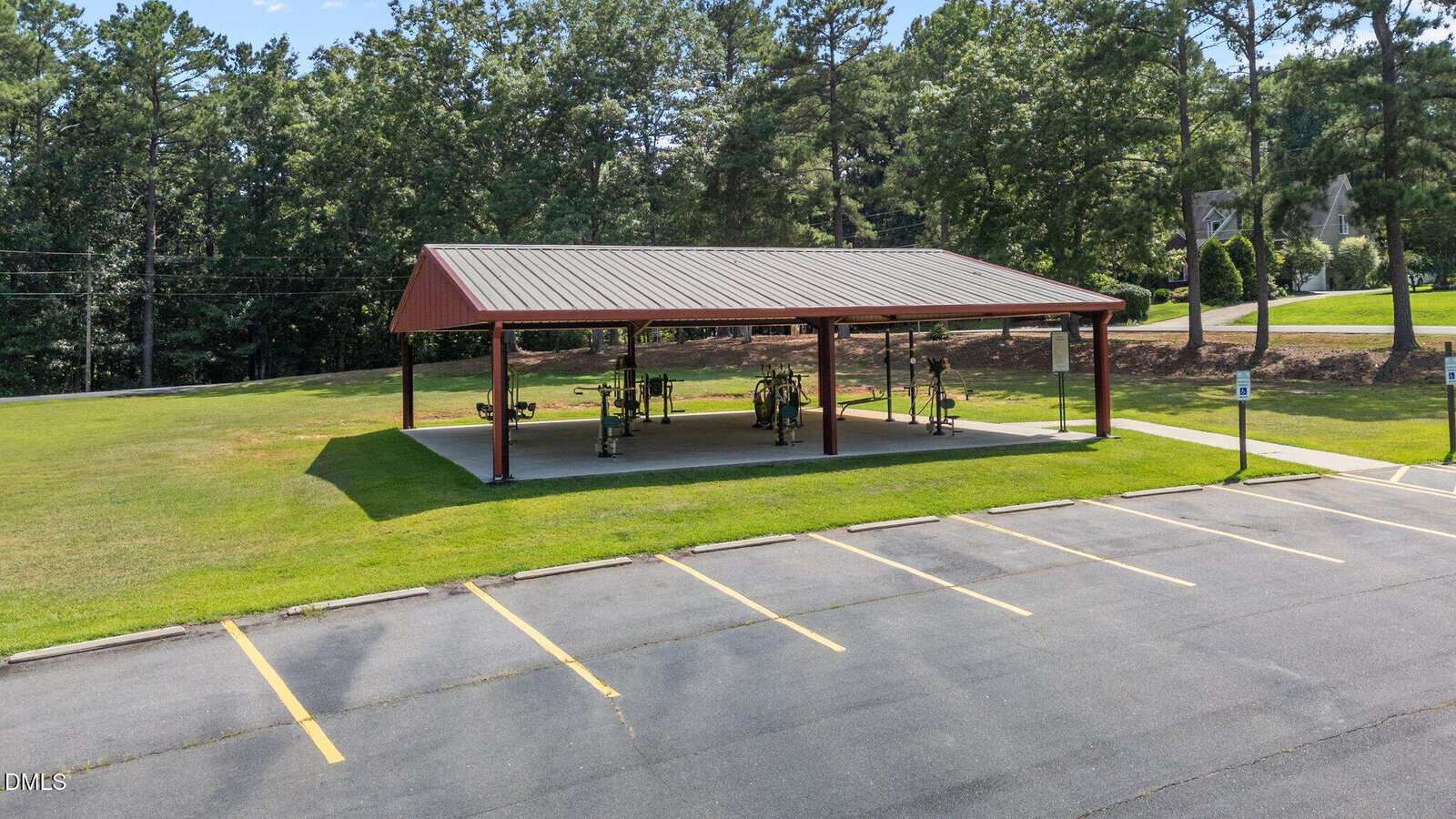 A covered outdoor exercise area with fitness equipment sits on a grassy lawn beside an empty parking lot, surrounded by trees and a residential area in the background.
