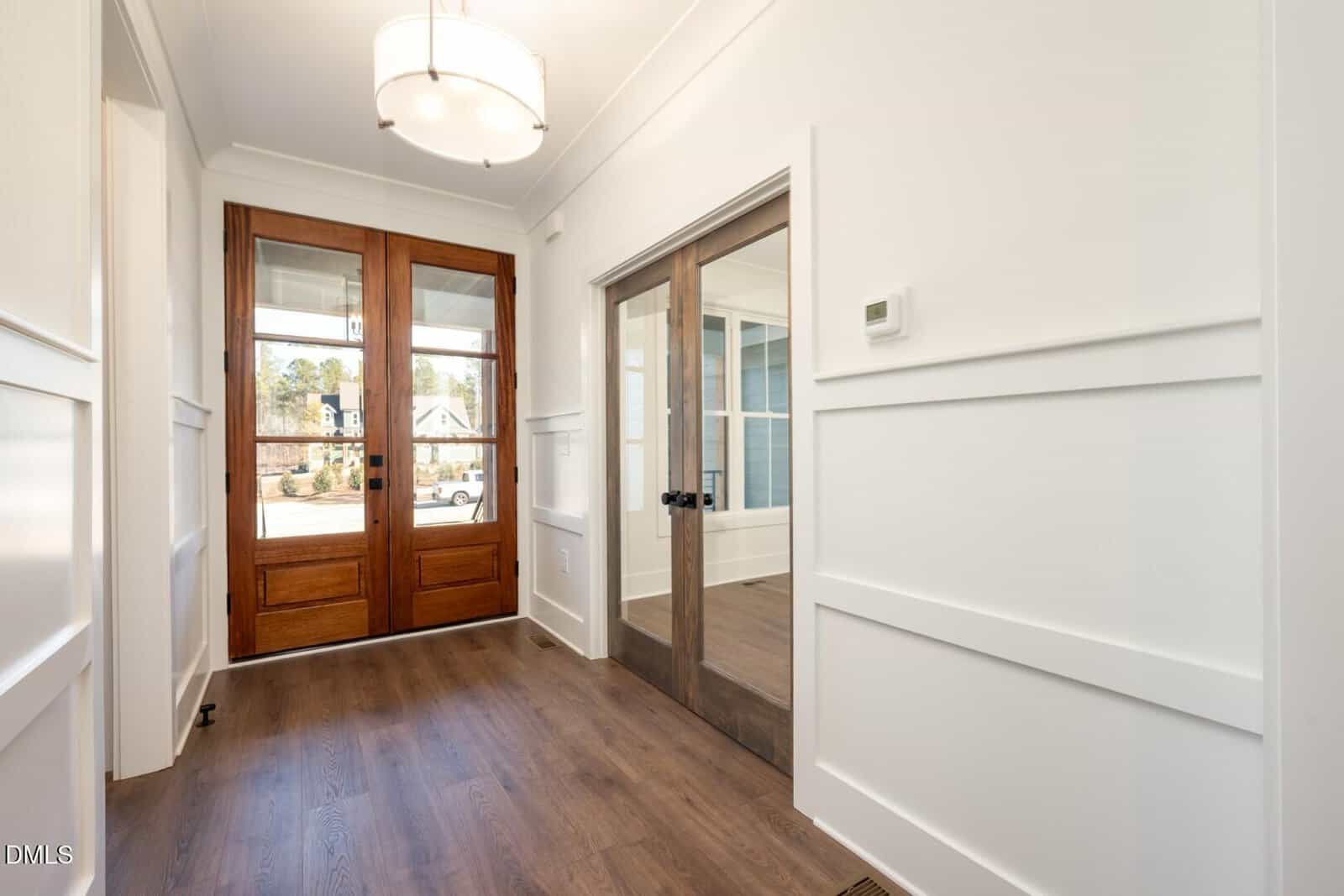 A bright entryway with wood flooring, white paneled walls, a double wooden front door with glass panels, a matching glass-paneled interior door, and a modern ceiling light fixture.