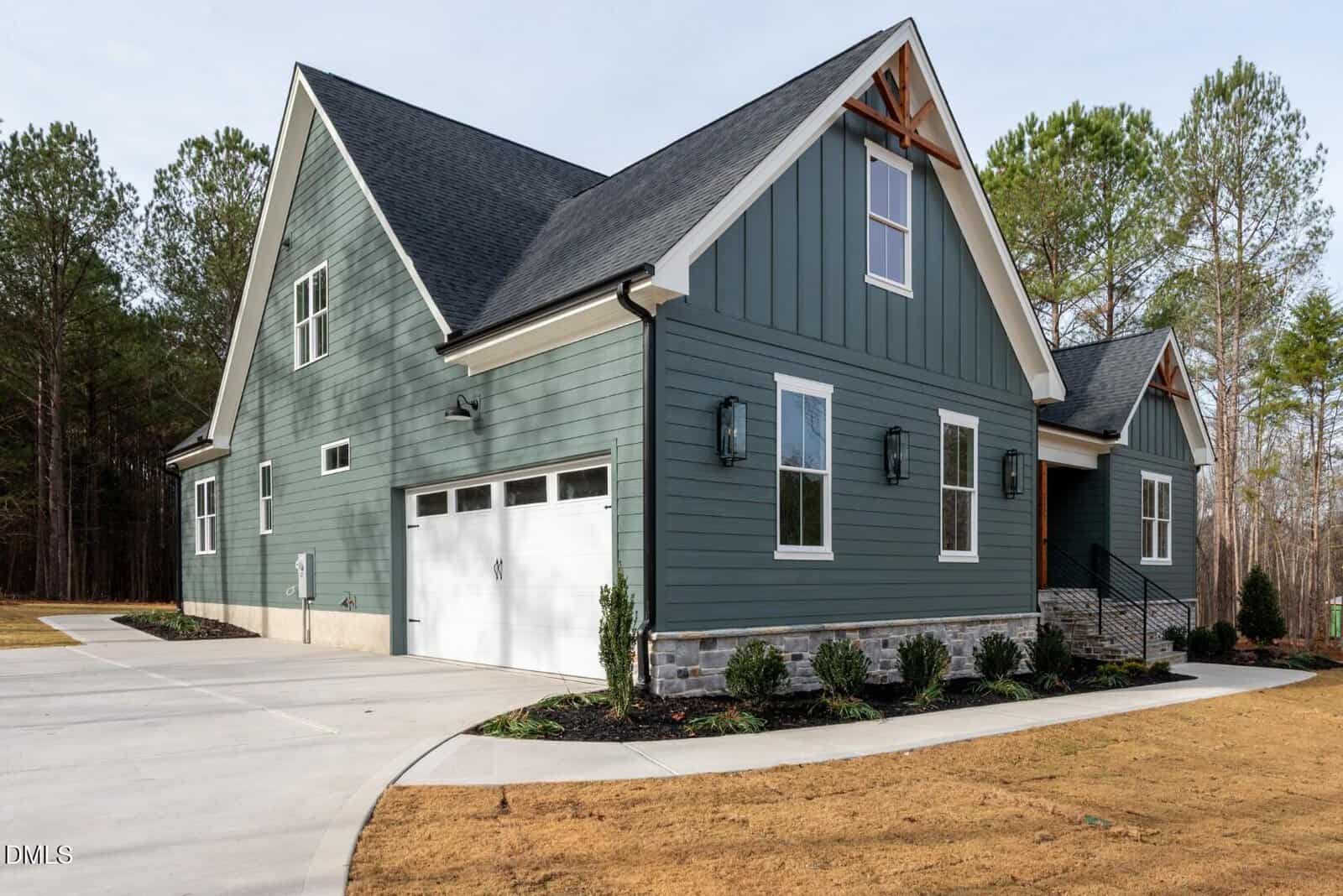 A modern two-story house with green siding, white trim, and a double garage. The driveway curves to the garage, and the landscaped yard features small shrubs. Tall trees are visible in the background.