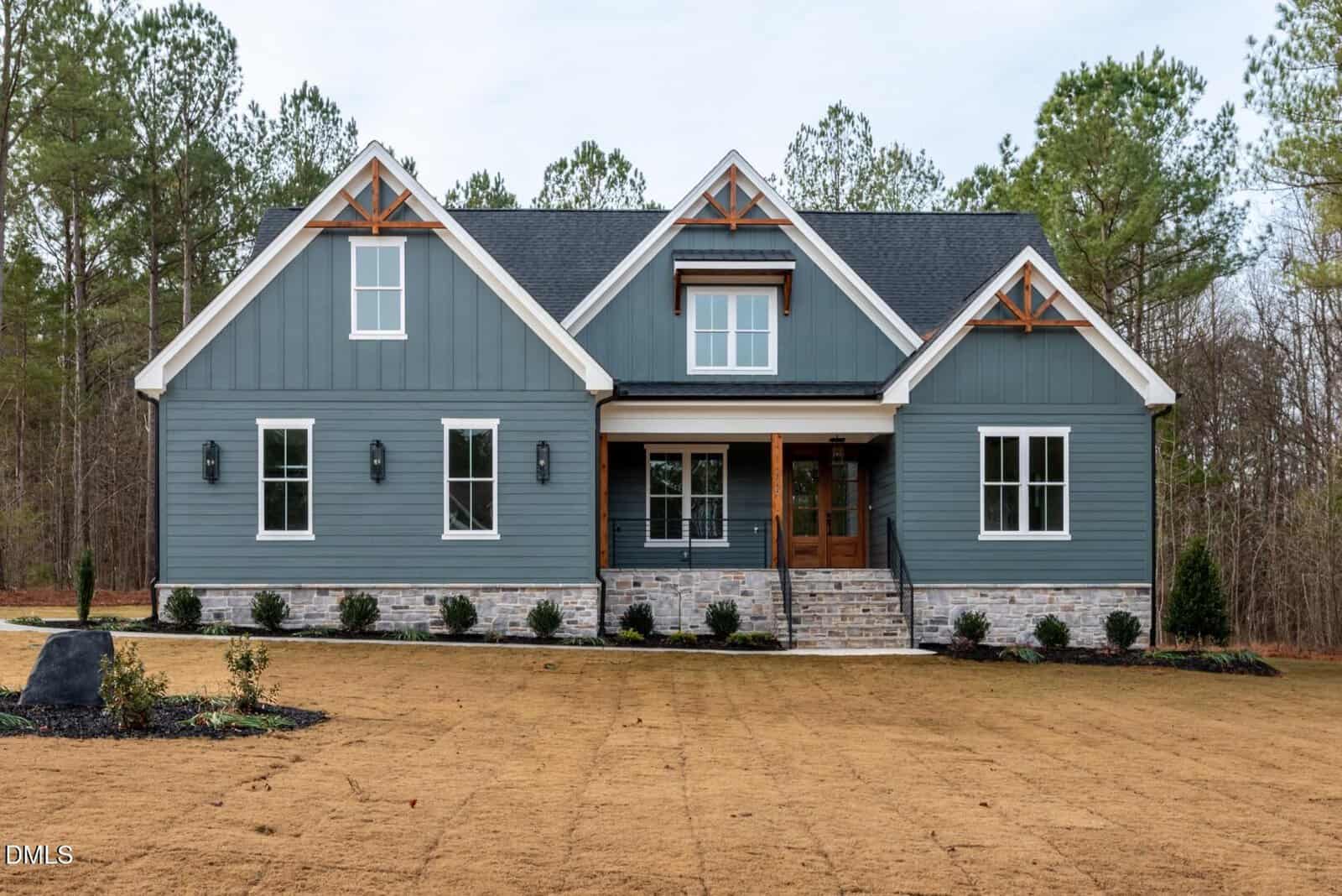 A modern blue-gray house with white trim, stone accents, and wooden gable details located at 275 Whistlers Cove in Louisburg. The home features a welcoming front porch with stairs and is surrounded by a brown lawn and trees in the background.