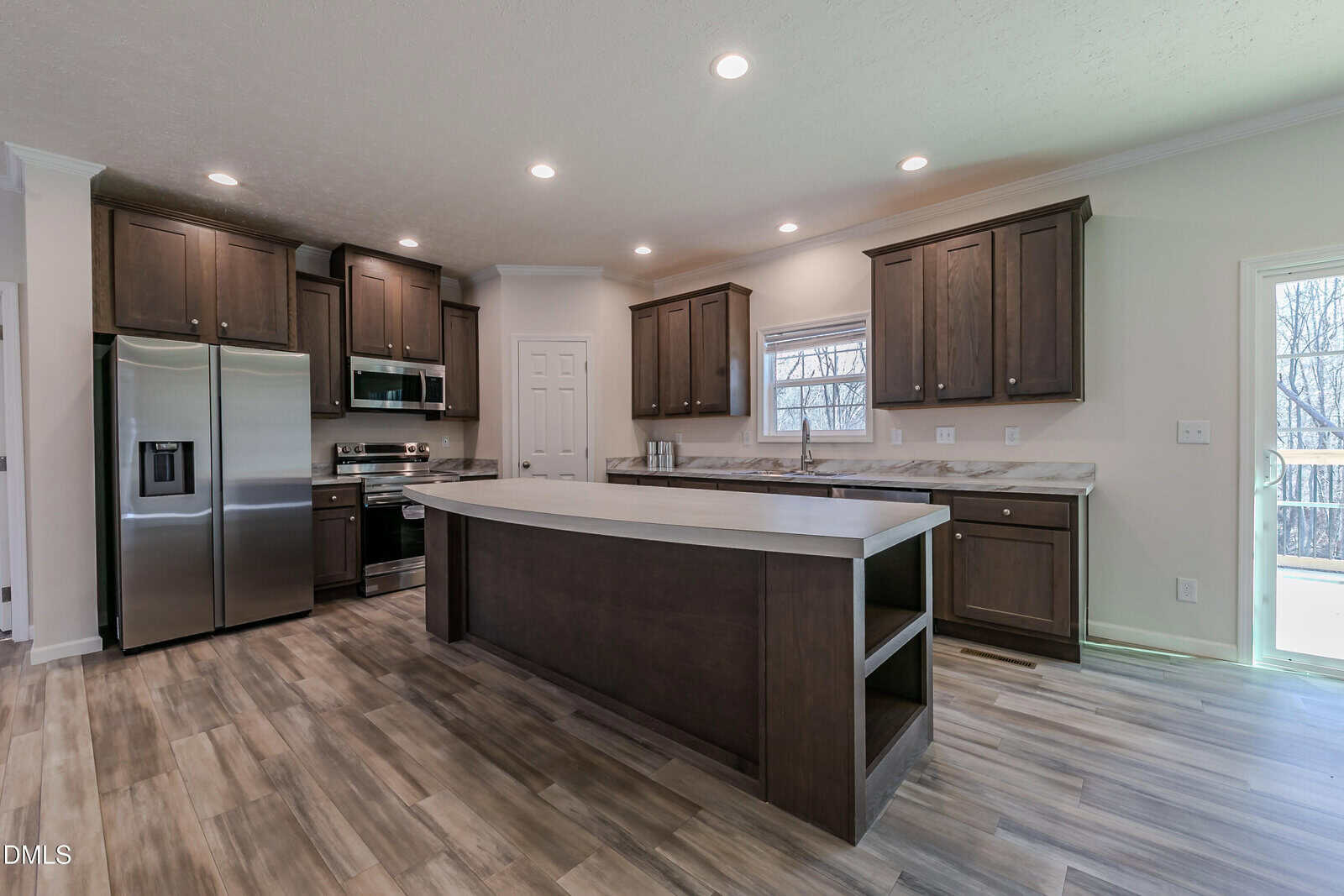 A modern kitchen with dark wood cabinets, stainless steel appliances, a large island with an overhang, marble countertops, and light wood flooring; recessed lighting and a window above the sink brighten the space.
