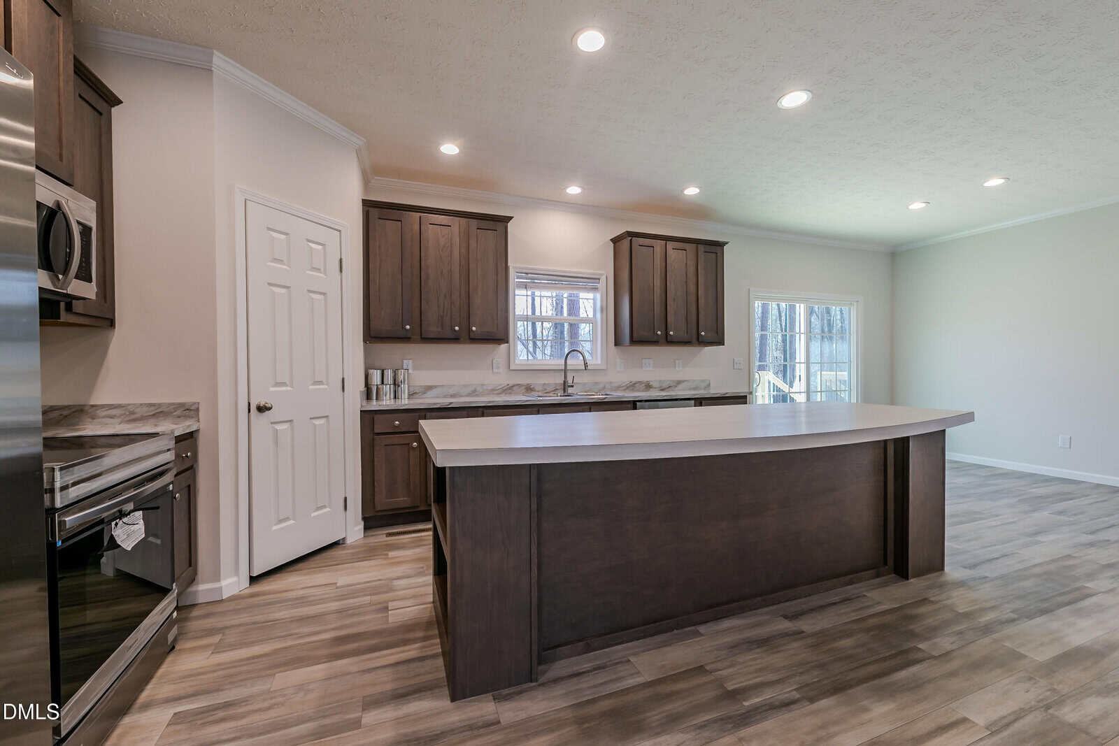 Modern kitchen with dark wood cabinets, a large island with a curved countertop, stainless steel appliances, wood-look flooring, and recessed lighting. A window and glass door provide natural light.