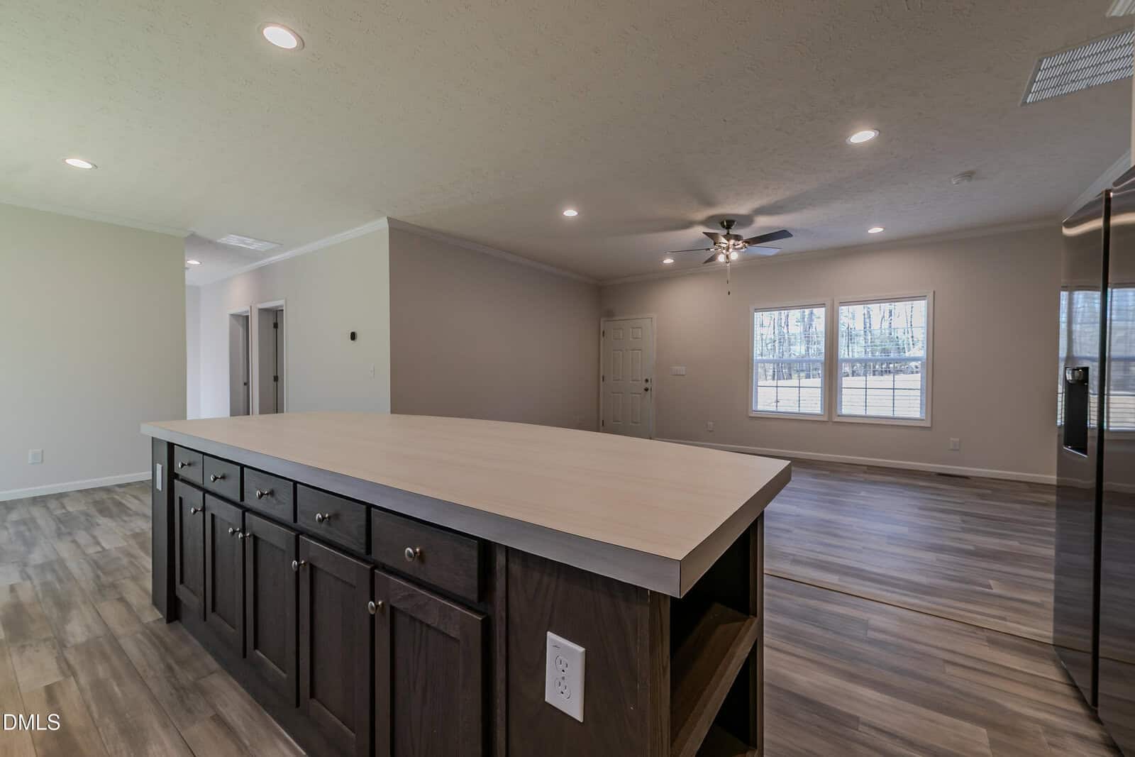 Open-concept living area with a large kitchen island, dark cabinets, wood flooring, beige walls, ceiling fan, and windows letting in natural light. Doorways lead to adjacent rooms.