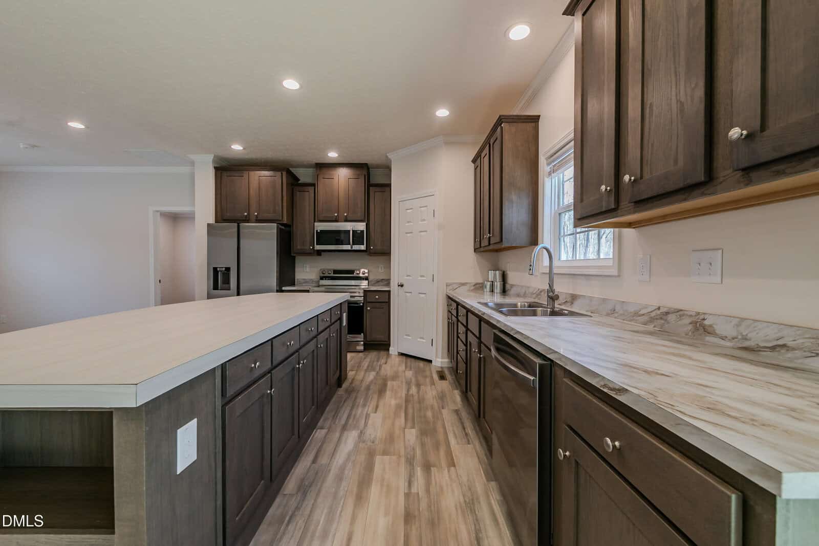 A spacious, modern kitchen with dark wood cabinets, marble countertops, and a large island. Stainless steel appliances, including a refrigerator and dishwasher, are visible.