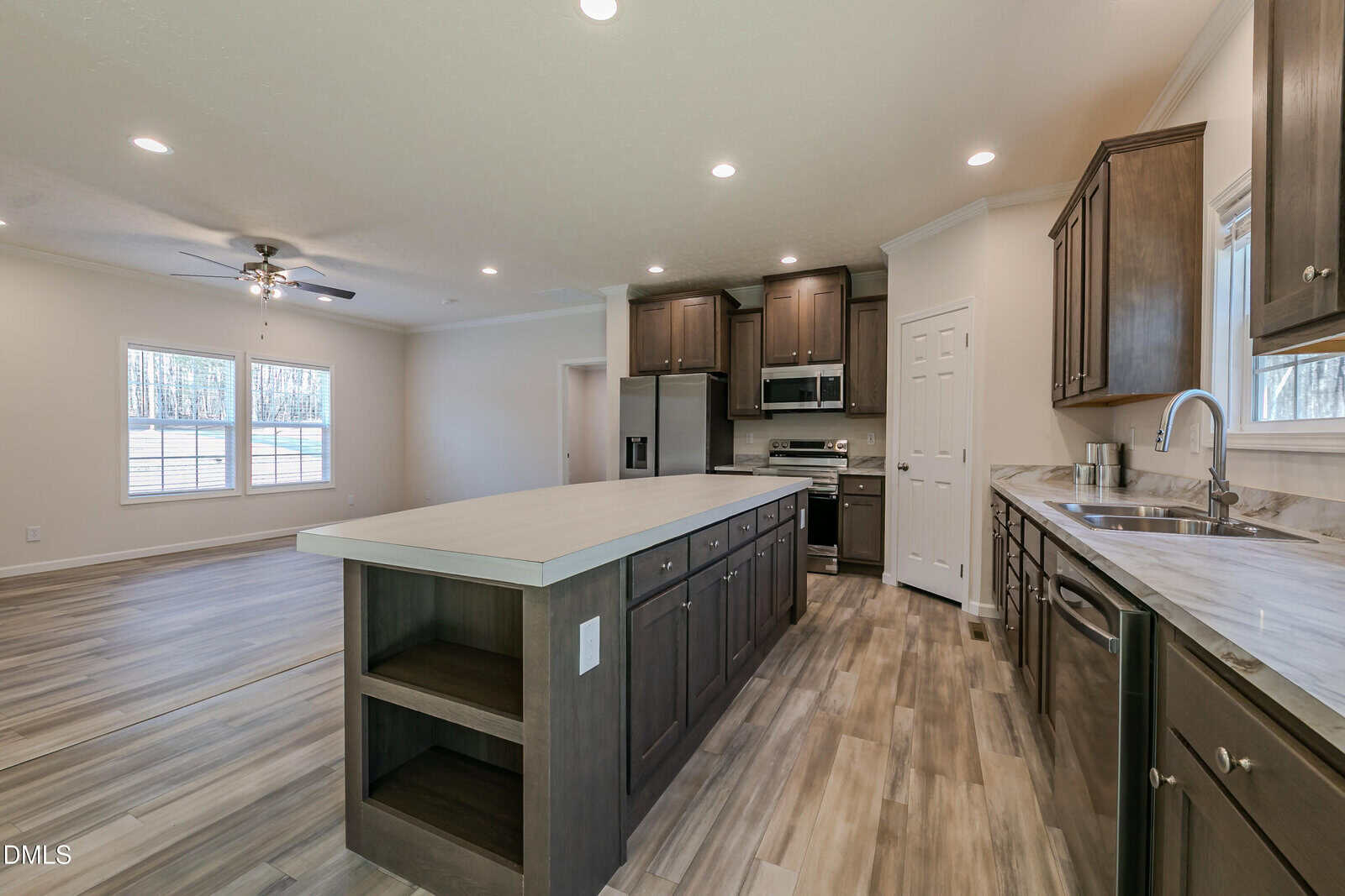 A modern kitchen features dark wood cabinets, a large island with shelves, stainless steel appliances, a double sink, and light wood plank flooring.