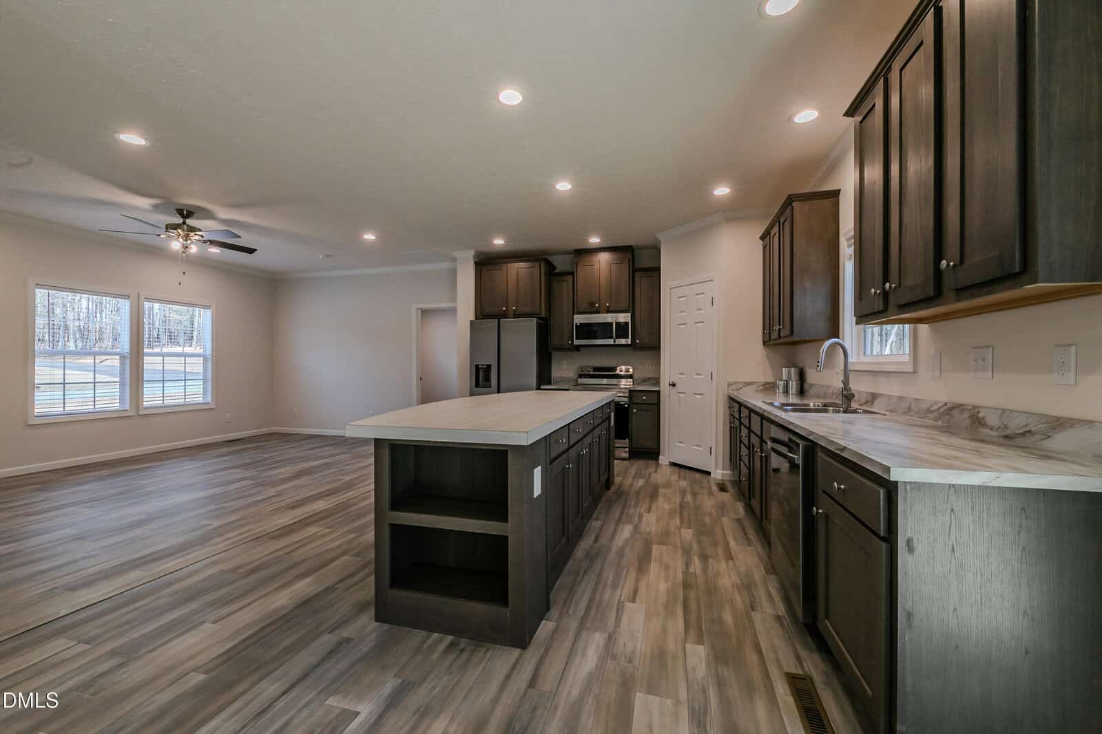 Modern open-concept kitchen with dark cabinets, light countertops, stainless steel appliances, and an island. Adjacent living area features large windows, wood-look flooring, and a ceiling fan.