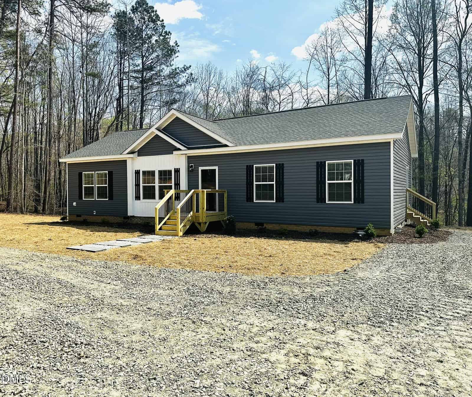 A modern single-story house with dark blue siding, white trim, and black shutters, sits in a wooded area. The home features a covered front porch with yellow railings and a gravel driveway in front.