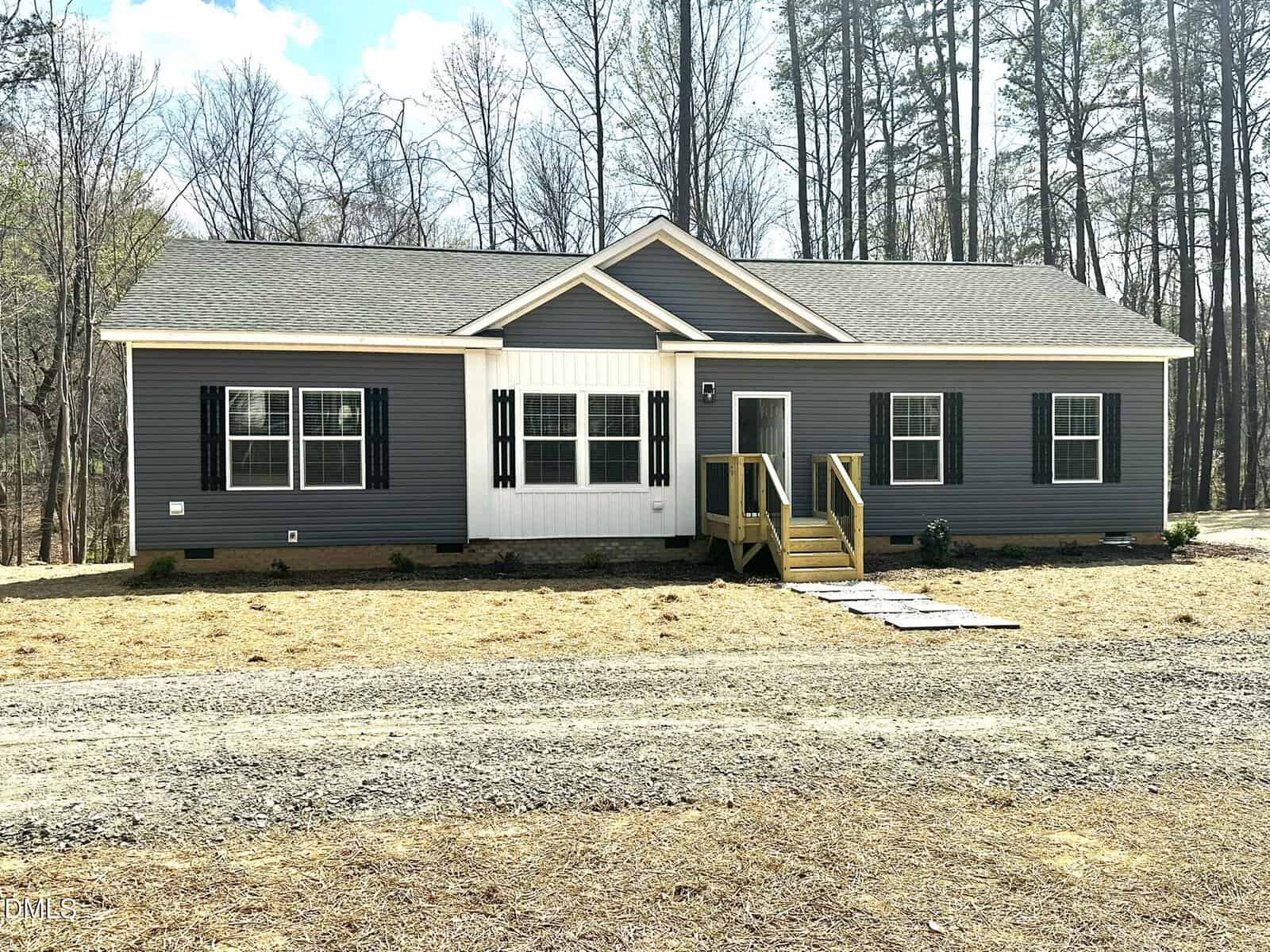 A single-story gray house with white trim, black shutters, and a small wooden front porch sits at 207 Old NC 75 S in Stem, nestled among bare trees with a dirt path leading to the entrance.