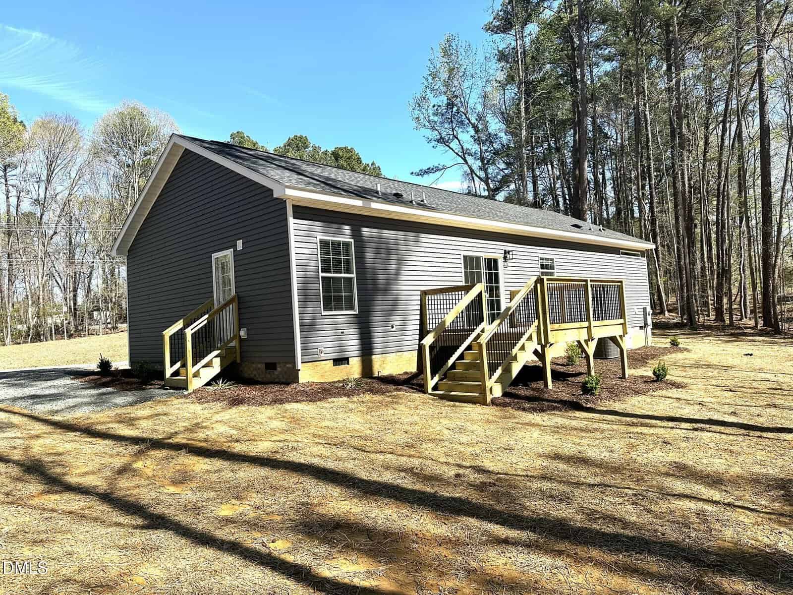 Single-story gray house with white trim, featuring a front porch with stairs and railings, set among tall trees and surrounded by a grassy, partially wooded yard under a clear blue sky.