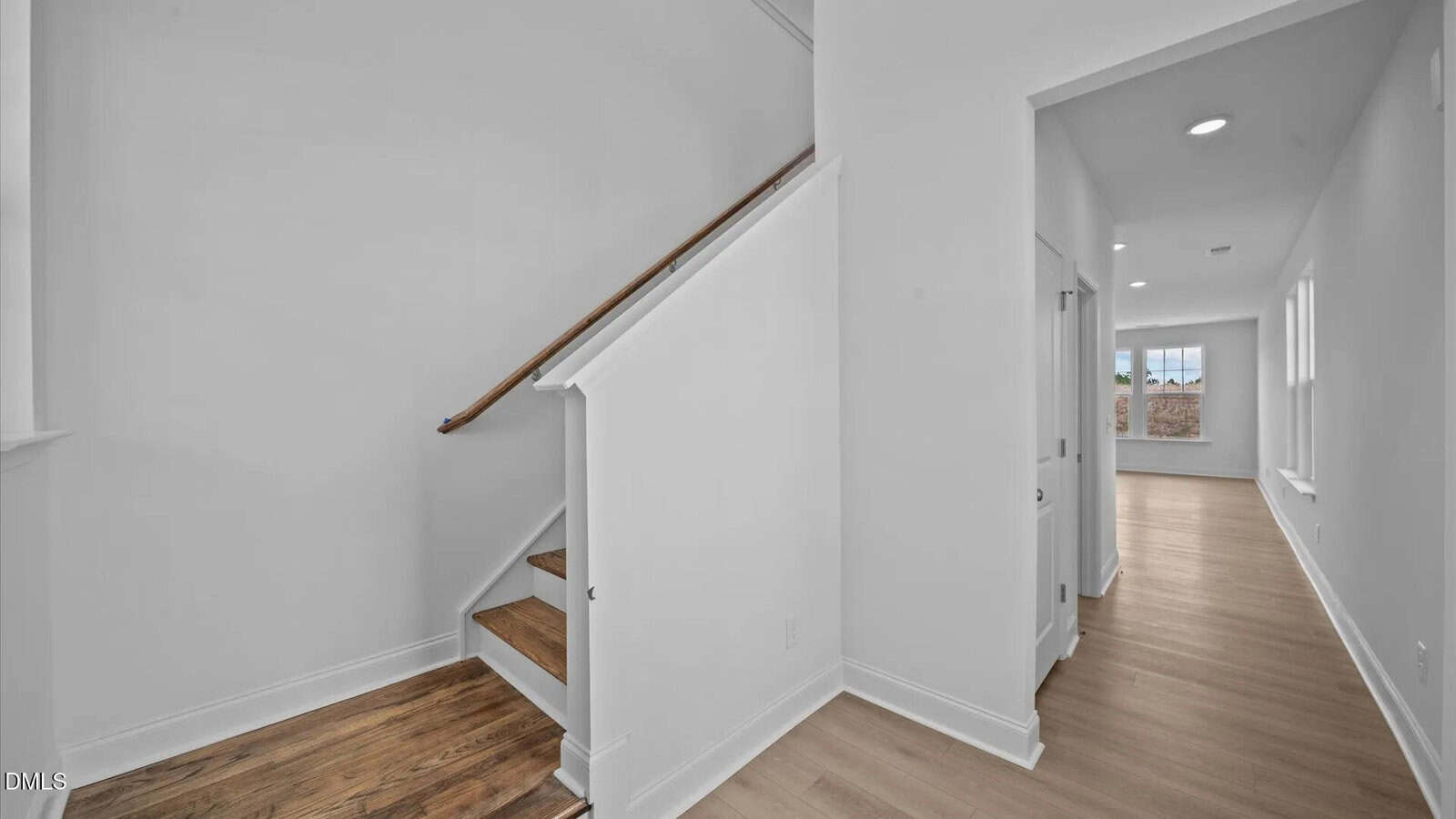 A bright hallway with light wood floors, white walls, and a wooden staircase with a simple handrail leading up. A corridor extends to a sunlit room with multiple windows in the background.