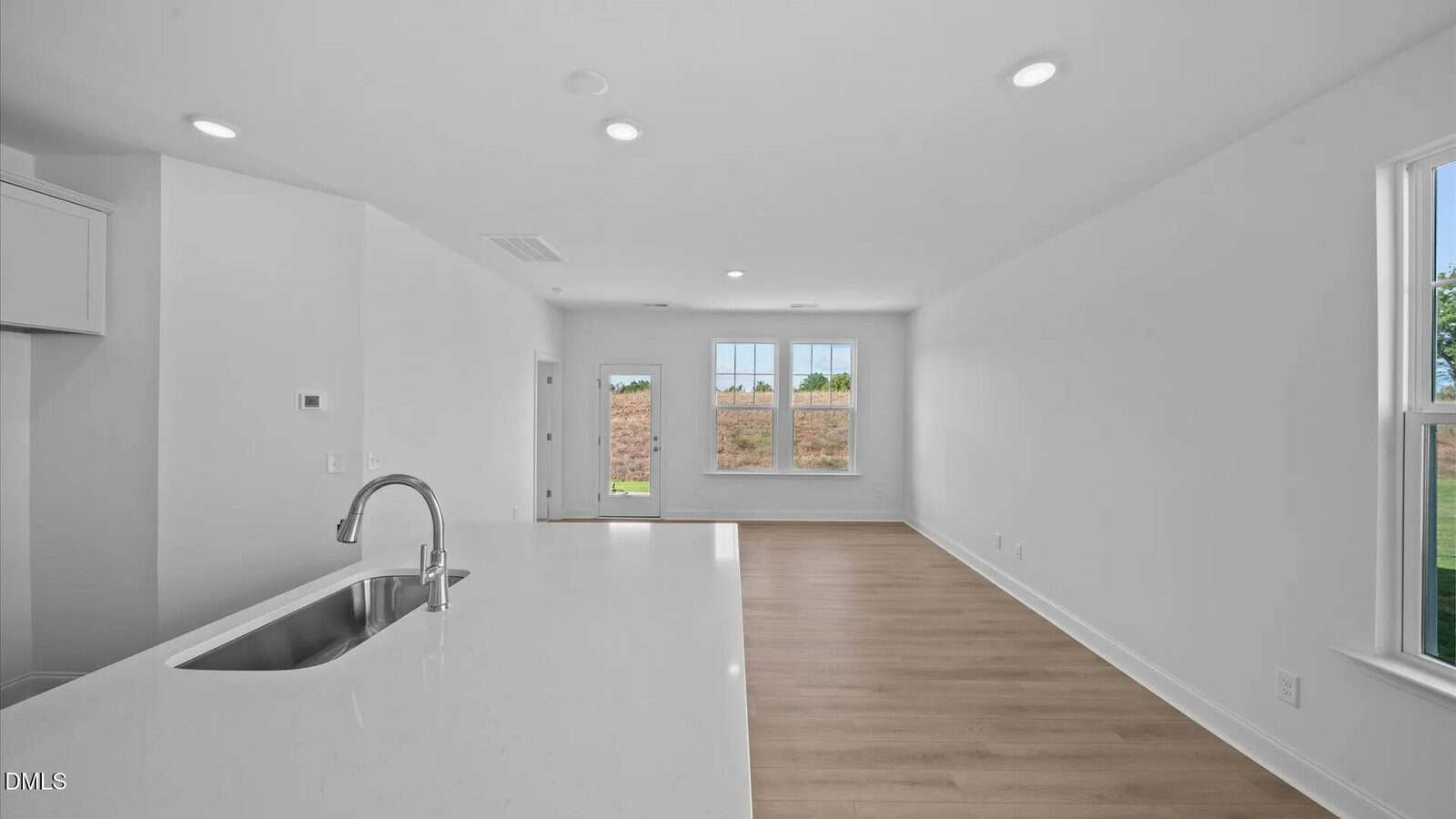 A modern, unfurnished open-concept living space with white walls, recessed lighting, large windows, and light wood flooring. A kitchen island with a sink is visible in the foreground on the left.
