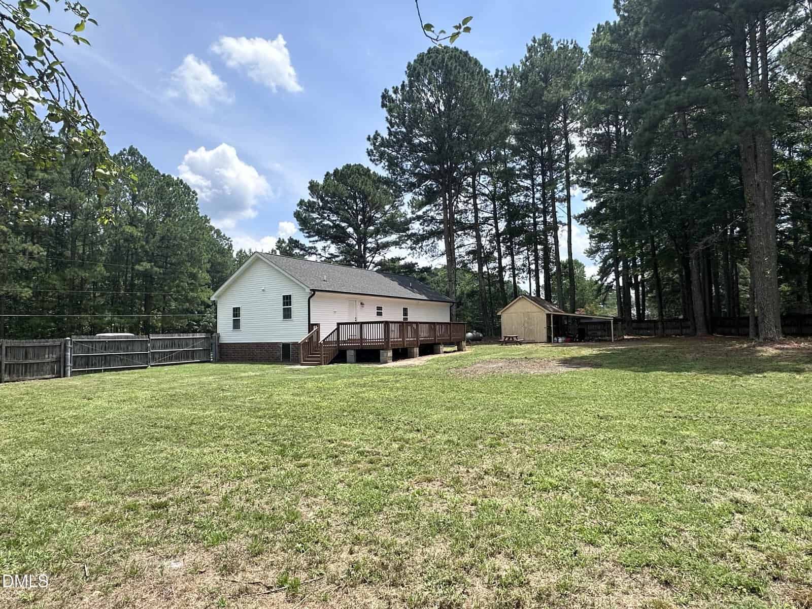 A white single-story house with a wooden deck is shown in a large, grassy backyard bordered by trees and a wooden fence. A small shed is visible near the edge of the wooded area under a partly cloudy sky.