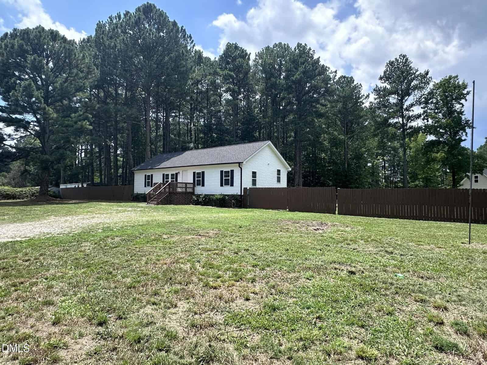A single-story white house with a dark roof sits at 96 Vineyard Drive, Louisburg, in front of a tall brown wooden fence and is surrounded by grass and trees. Steps lead to the front door, with a wooded area visible behind the house.
