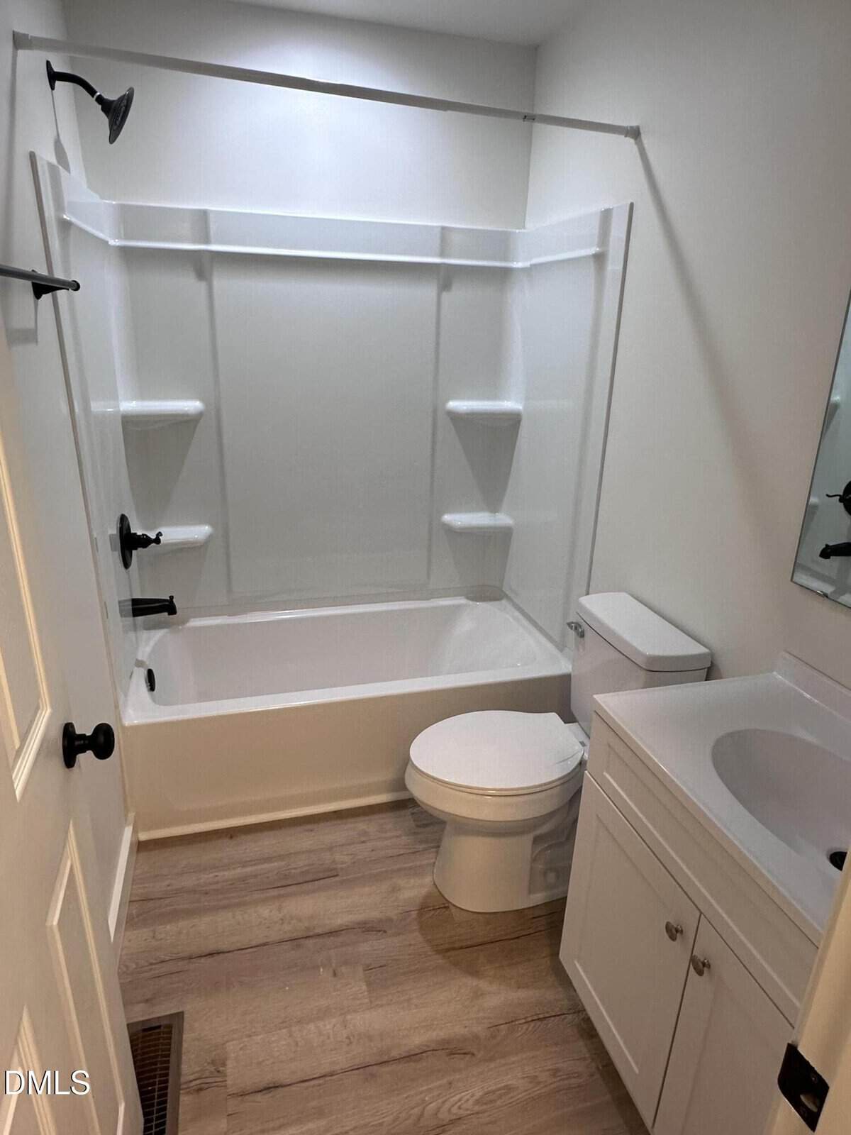 A small bathroom with a white bathtub-shower combo, white toilet, and white vanity with sink and mirror. The floor has light wood-style planks and the walls are painted white.