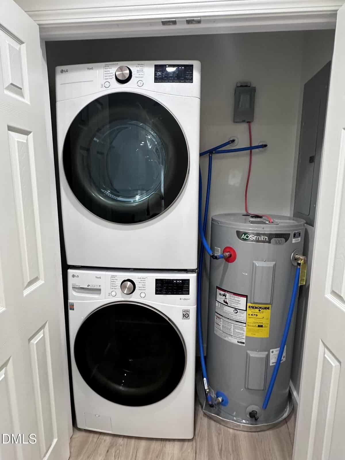 A small laundry closet with a stacked white front-load washer and dryer on the left and a gray AO Smith water heater with attached hoses on the right. The floor is laminate wood, and white doors frame the closet.