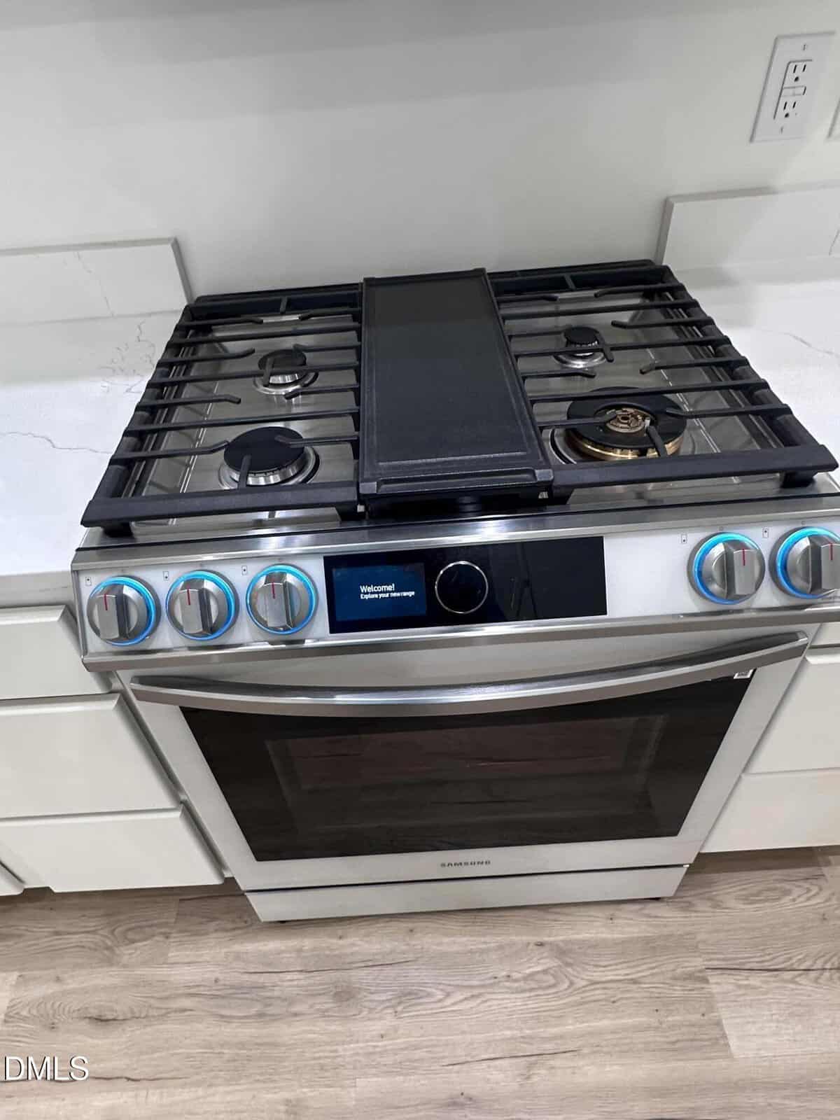 A stainless steel Samsung gas stove and oven with four burners and a center griddle, surrounded by white countertops and cabinetry, is shown in a modern kitchen.