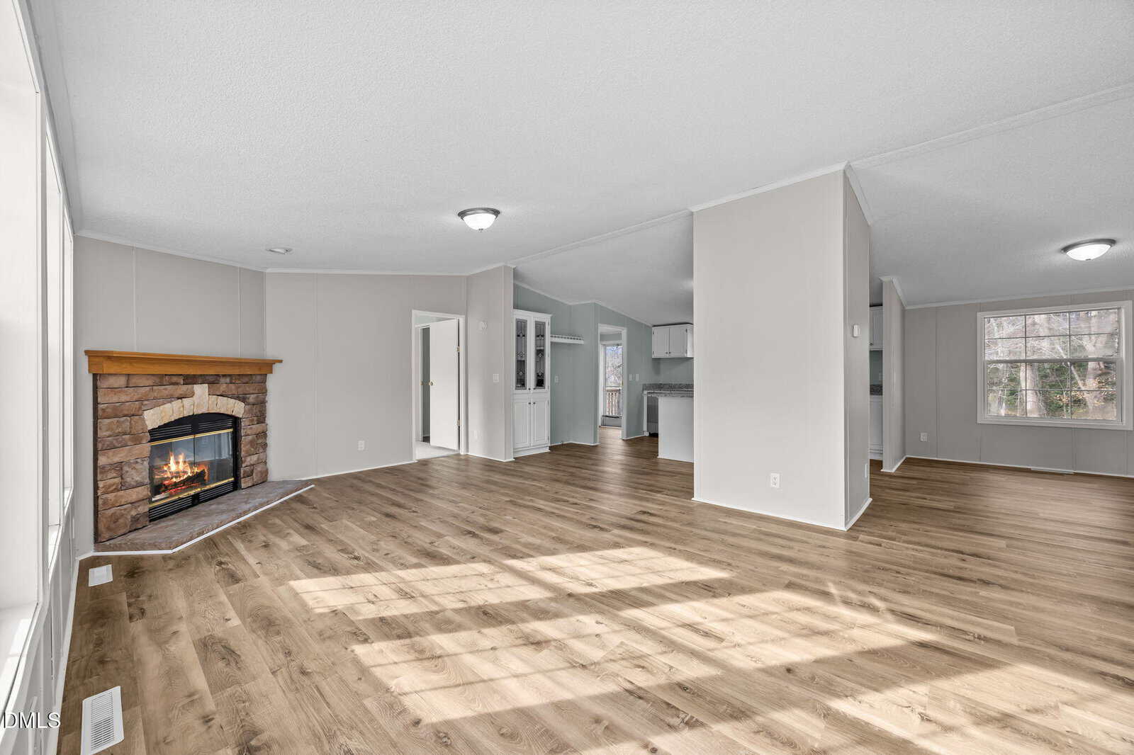Spacious living room with light wood flooring, a corner stone fireplace, and neutral-colored walls. Large windows provide natural light. Open layout leads to other rooms, with a built-in cabinet visible in the background.