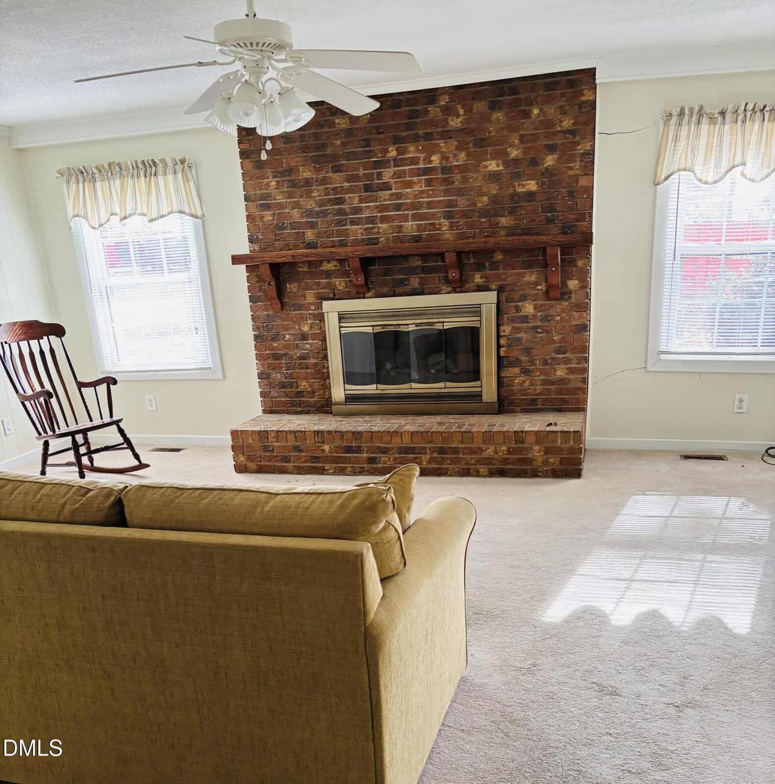 A living room with a brick fireplace, a brass-trimmed fireplace insert, a beige couch facing the fireplace, a wooden rocking chair, two windows with curtains, cream walls, and a white ceiling fan.