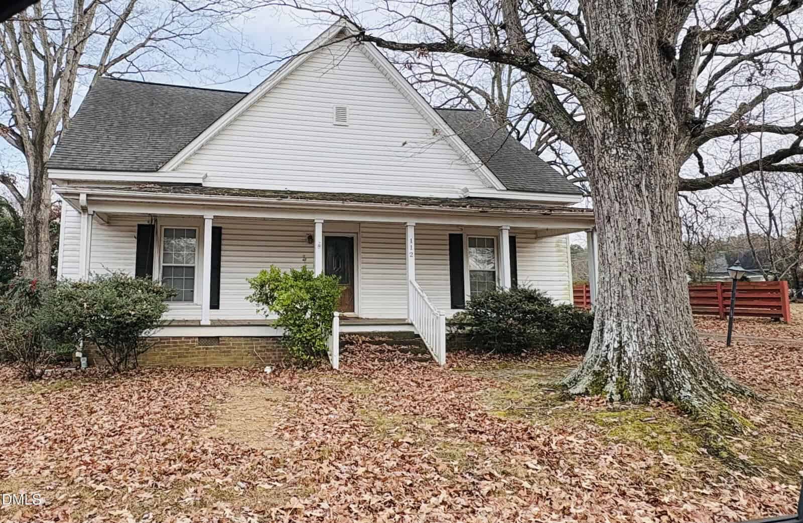 Nestled at 112 N Nassau Street in Youngsville, this white, single-story house features a welcoming covered front porch, accented by classic black shutters and two front windows. A large tree provides shade over the yard, where fallen leaves blanket the ground and walkway. Bushes border the porch, while a red fence adds charm on the right side of the property.