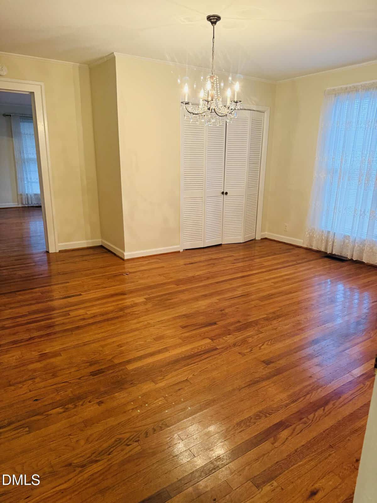 Empty room with polished wooden floor, cream-colored walls, a white closet with double doors, a window with lace curtains, a ceiling chandelier, and two doorways leading to adjacent rooms.