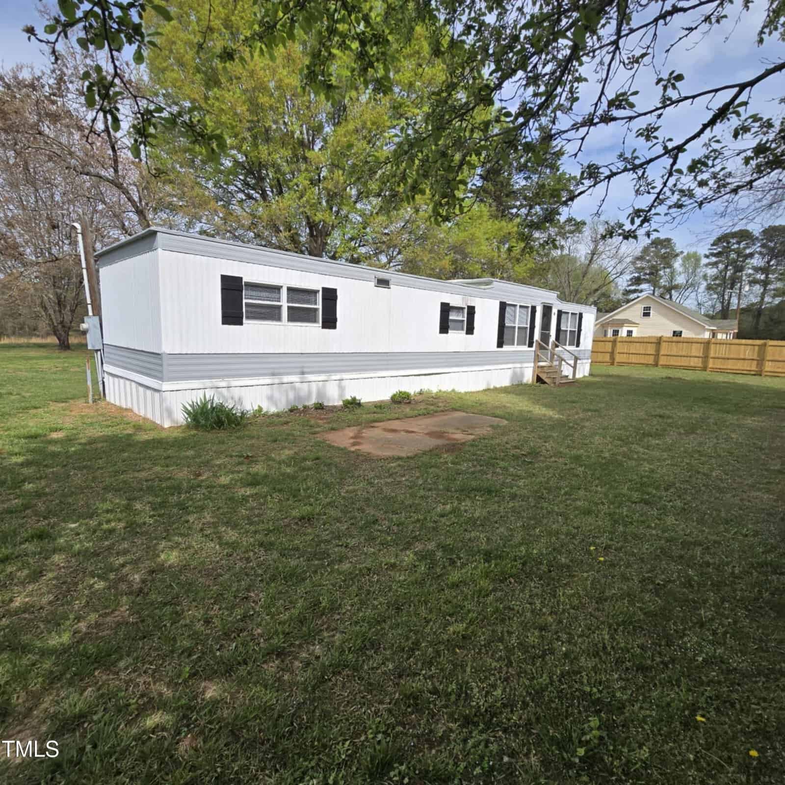 A white mobile home with black shutters sits on a grassy yard, bordered by trees and a wooden fence. The home has multiple windows and a small raised entrance with steps.