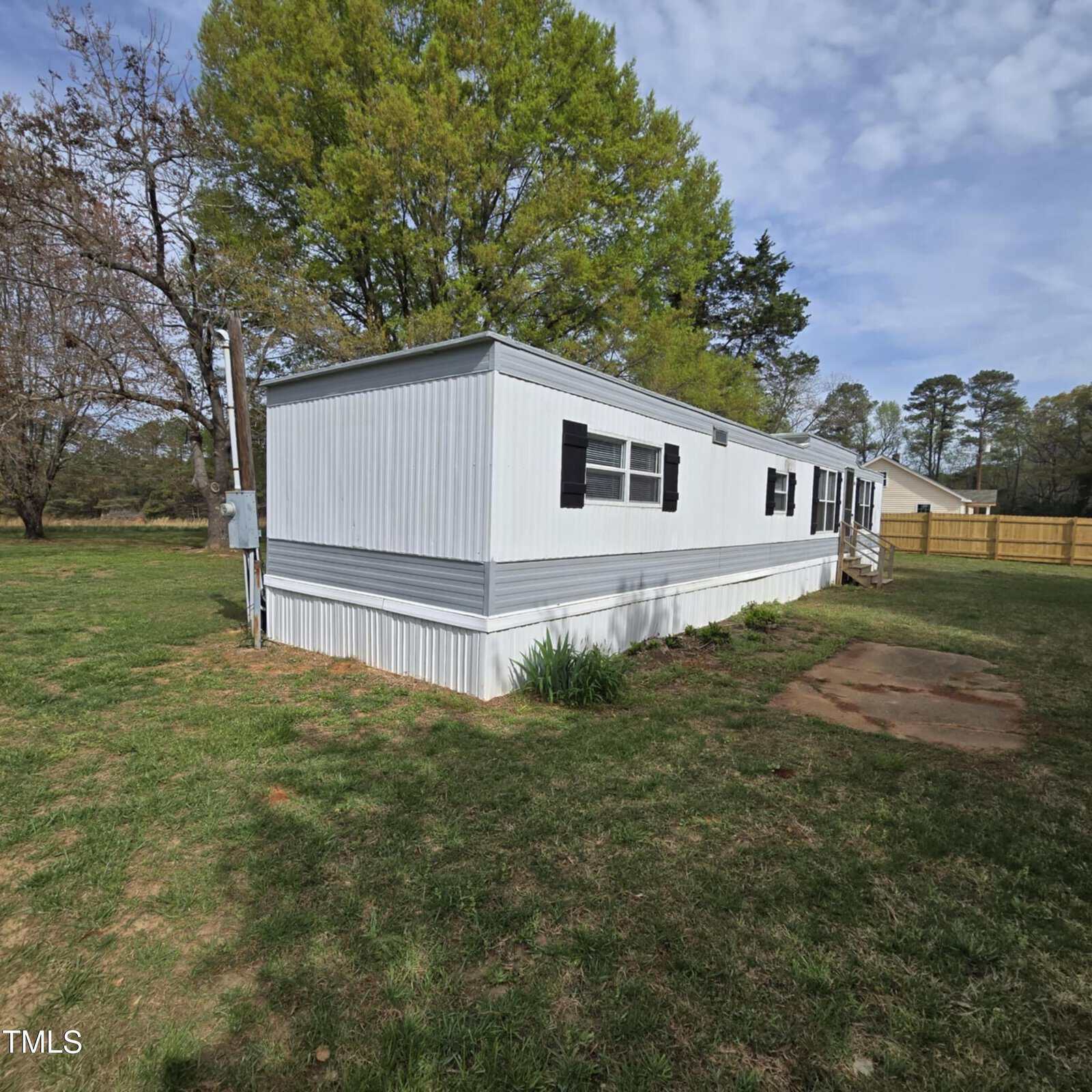A white mobile home with black shutters sits on a grassy lot with a few trees nearby, under a partly cloudy sky. A small concrete pad is visible in the foreground.