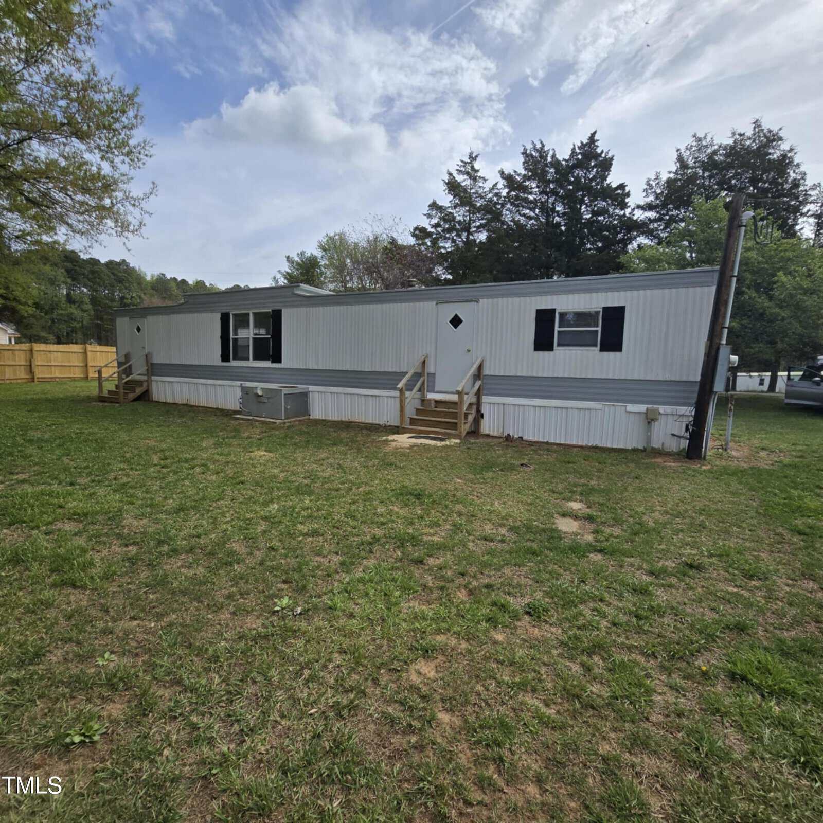 A single-wide mobile home with white siding and black shutters sits on a grassy yard. There are two small wooden porches with steps, and trees and a wooden fence are visible in the background.