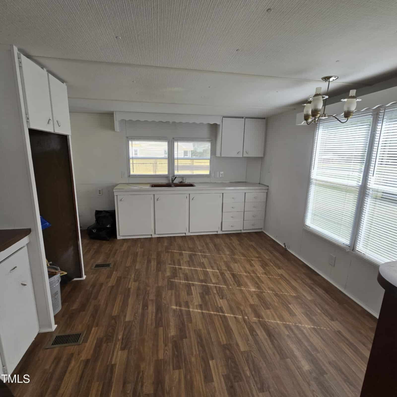 A vacant kitchen with wood-patterned vinyl flooring, white cabinets and drawers, a sink under two windows, a ceiling light fixture, and large windows letting in natural light.