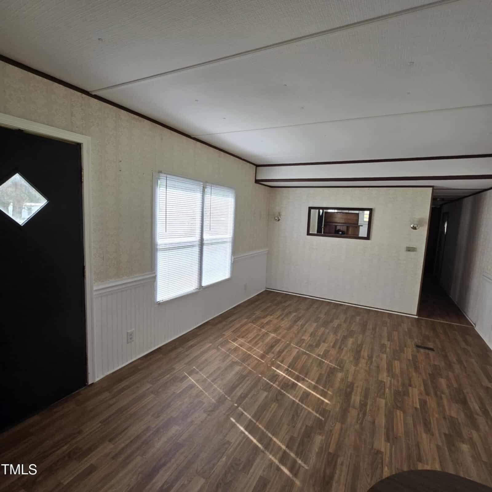 A bright, empty room with wood-look flooring, beige wallpaper, a black door with a diamond-shaped window, two windows letting in sunlight, a wall mirror, and a hallway leading to the back.
