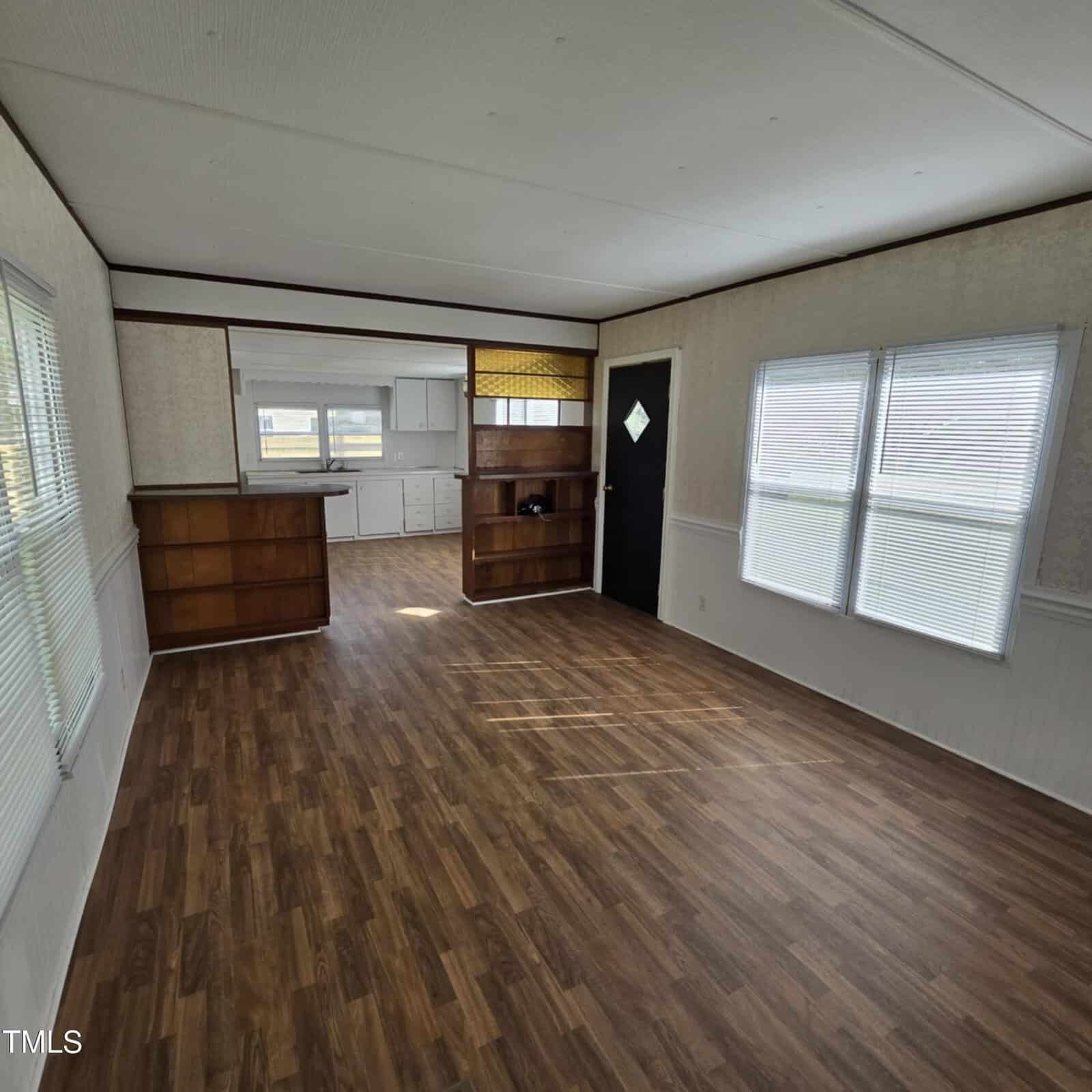 A spacious, empty room with wood flooring, large windows letting in natural light, and an open kitchen area with cabinets and shelving along one wall. The front door is black with a diamond-shaped window.