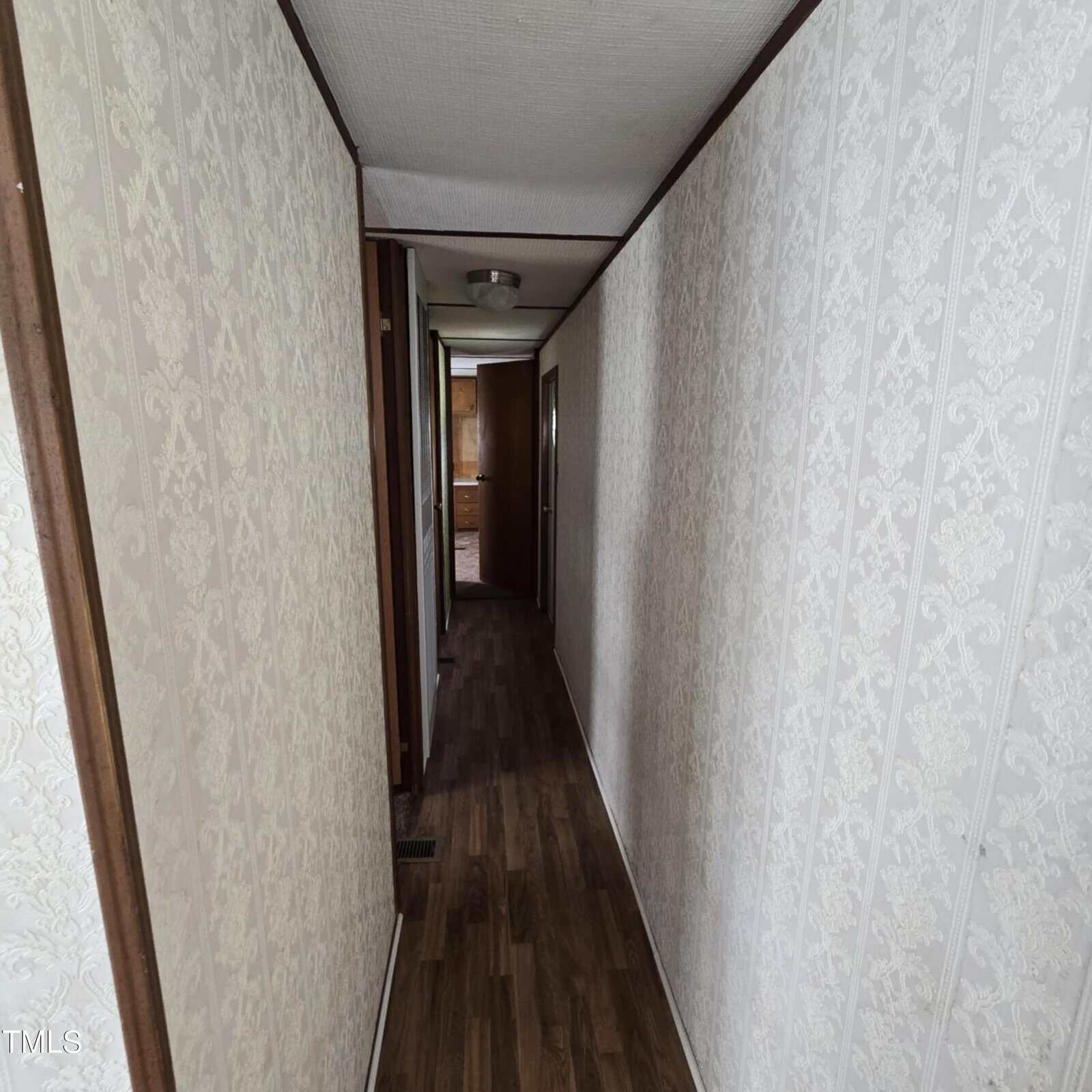 A narrow hallway with patterned wallpaper and wood flooring, leading to rooms at the end. The ceiling has two light fixtures, and doors line the sides of the hallway.