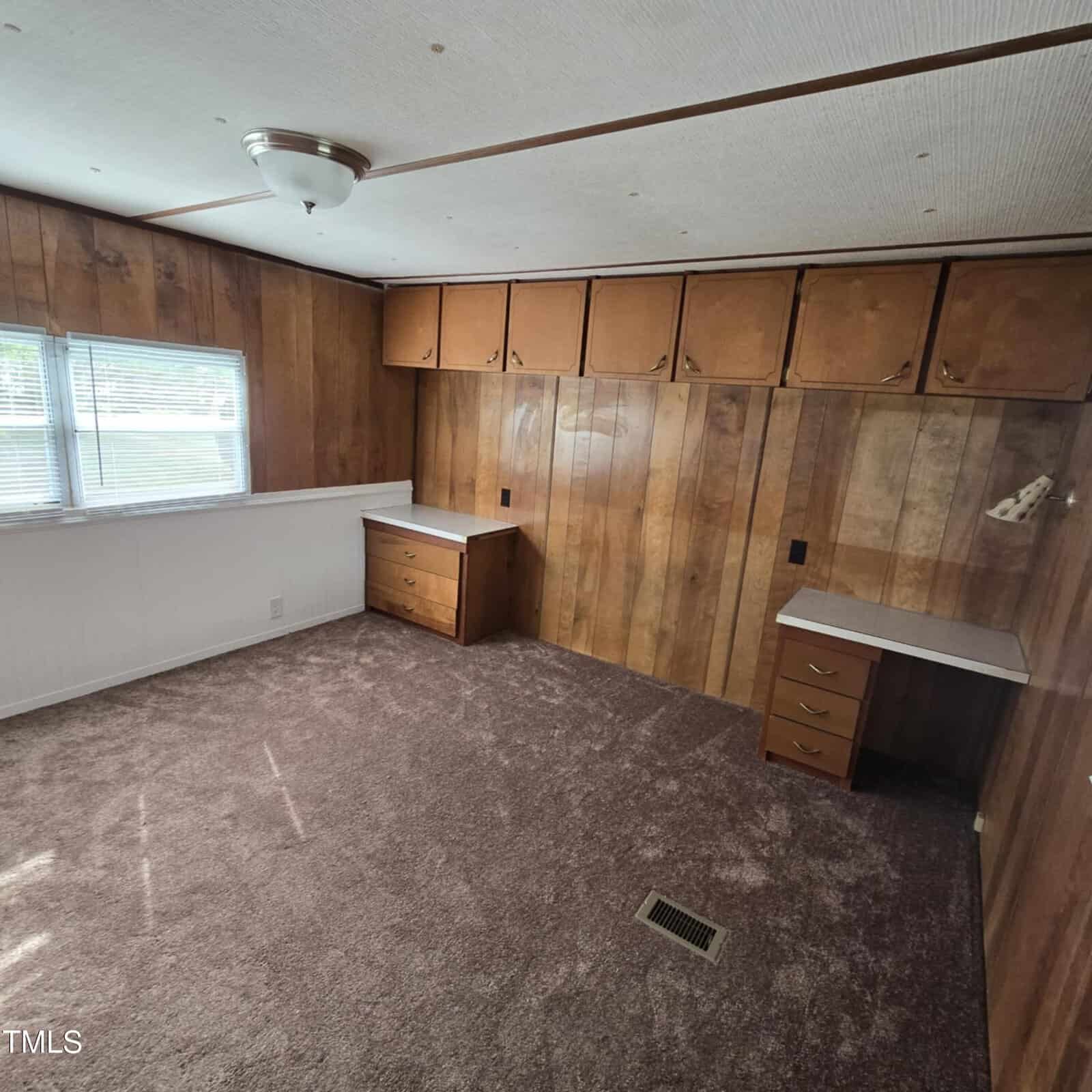 A room with wood-paneled walls, brown carpet, two built-in desks with drawers, overhead cabinets, a ceiling light, and a window with white blinds letting in natural light.