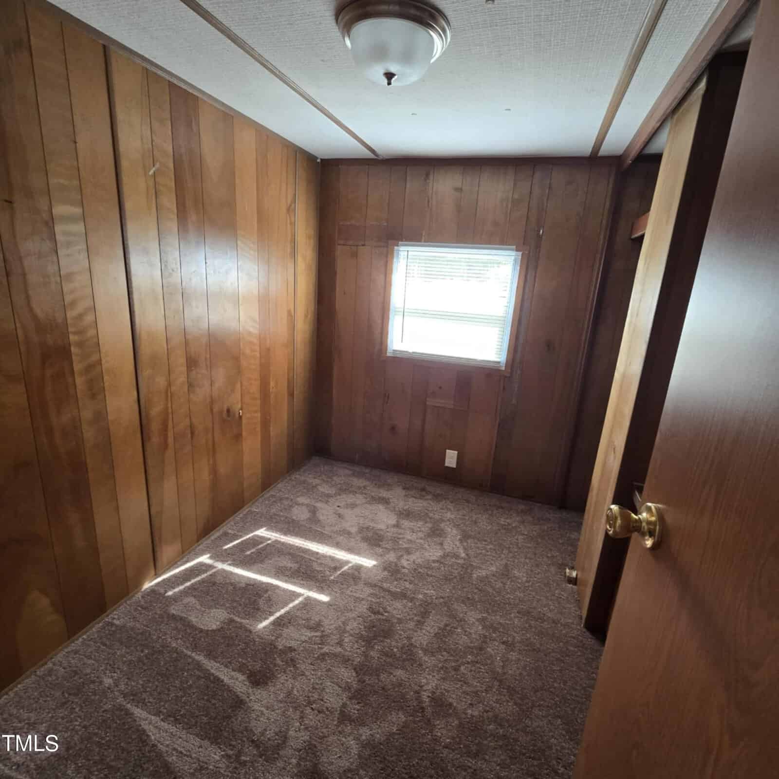 Small room with wood-paneled walls, brown carpeted floor, a single window with blinds letting in sunlight, a ceiling light fixture, and a partially open wooden door with a brass knob.