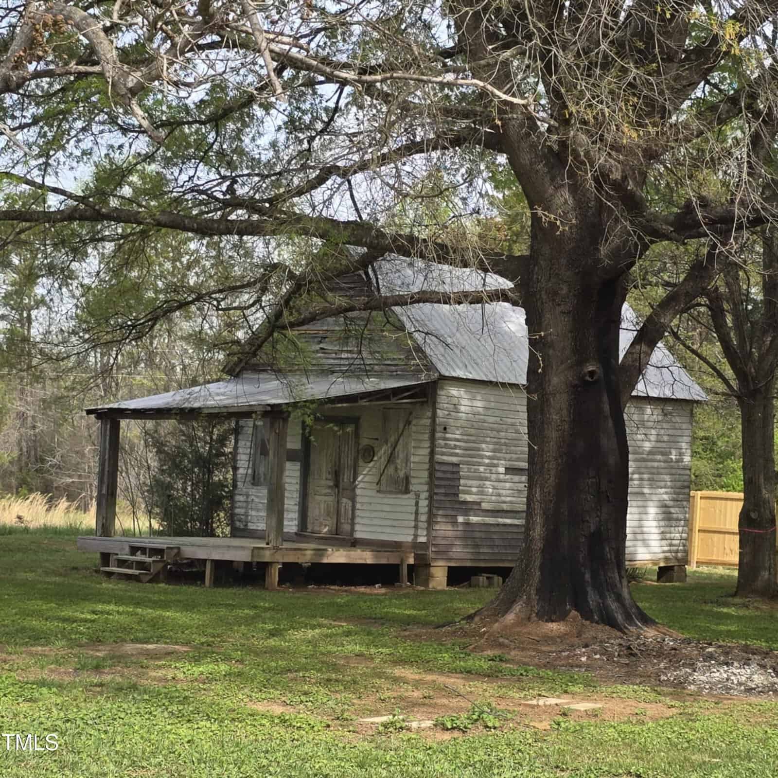 An old, weathered wooden house with a tin roof sits on a grassy lawn under large trees, with a wooden fence and wooded area in the background.