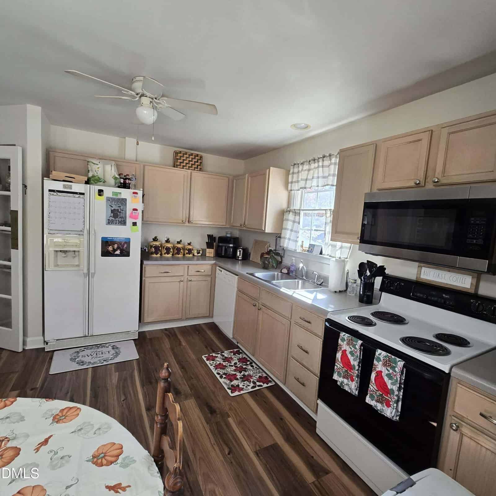 A kitchen with light wood cabinets, white appliances, a double oven with holiday-themed towels, a refrigerator with magnets and notes, a coffee maker, and a table with a pumpkin-patterned tablecloth.