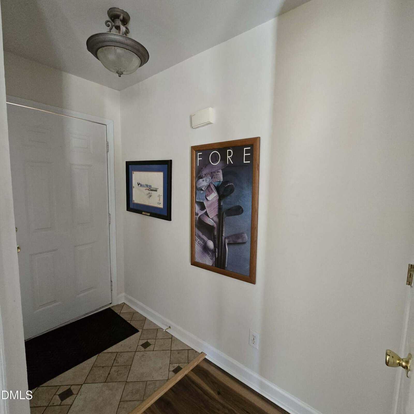 A small hallway with light-colored walls, a tiled and wood floor, and a white door. Two framed pictures hang on the wall, one with golf clubs and the word "FORE." A ceiling light fixture is visible above.