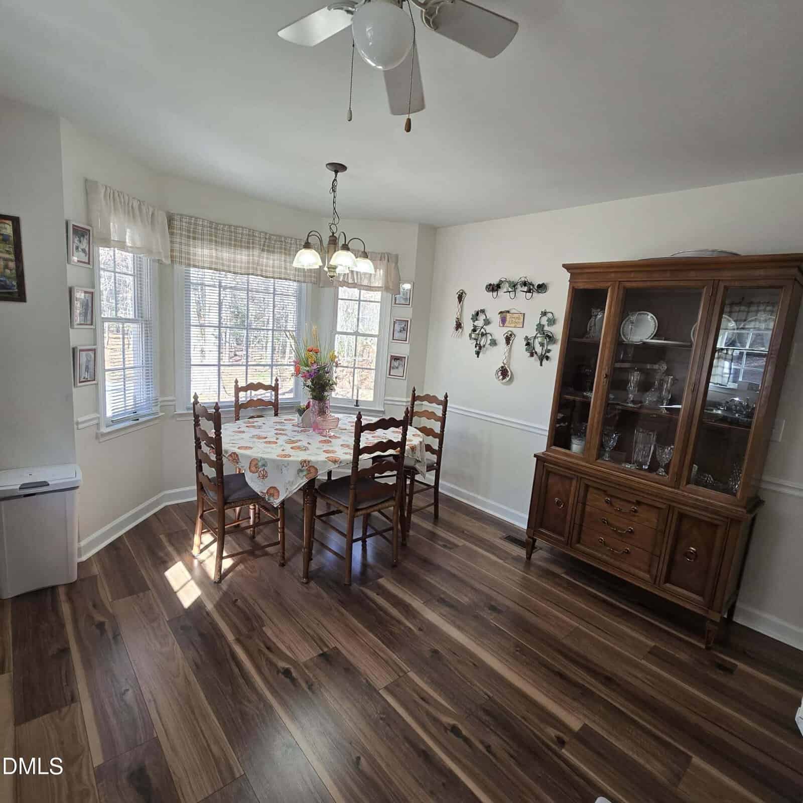 A dining room with wood flooring, a rectangular table covered by a floral tablecloth, four wooden chairs, a bay window with curtains, a ceiling fan, a chandelier, and a china cabinet with glass doors.