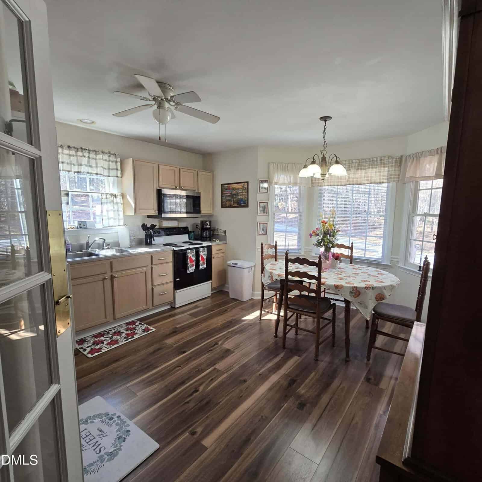 A kitchen with light wood cabinets, black appliances, a ceiling fan, and a dining area with a round table and four chairs. Large windows let in natural light, and the floor is a dark wood laminate.
