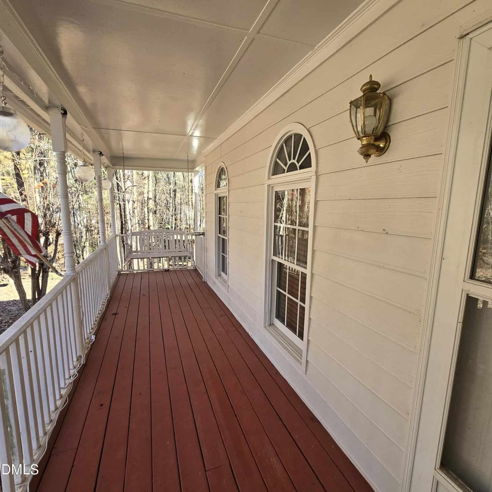A covered porch with red wooden flooring, white railing, and beige siding. Two large arched windows and a brass wall-mounted light fixture are visible. An American flag hangs on the left side. Trees can be seen in the background.