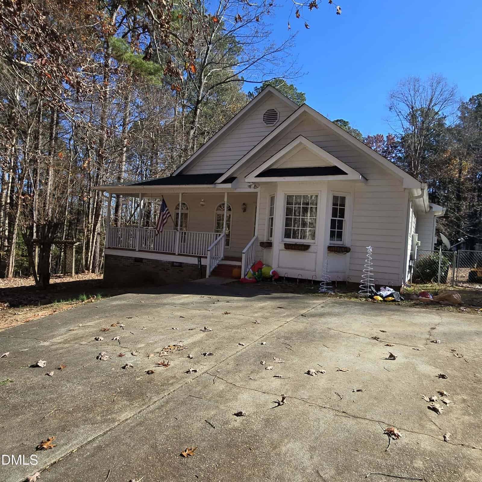 A single-story white house with a front porch and gable roof sits next to a wooded area at 122 Buckskin Drive, Louisburg. The wide, cracked driveway leads to a yard where toys and a small American flag are visible near the porch.