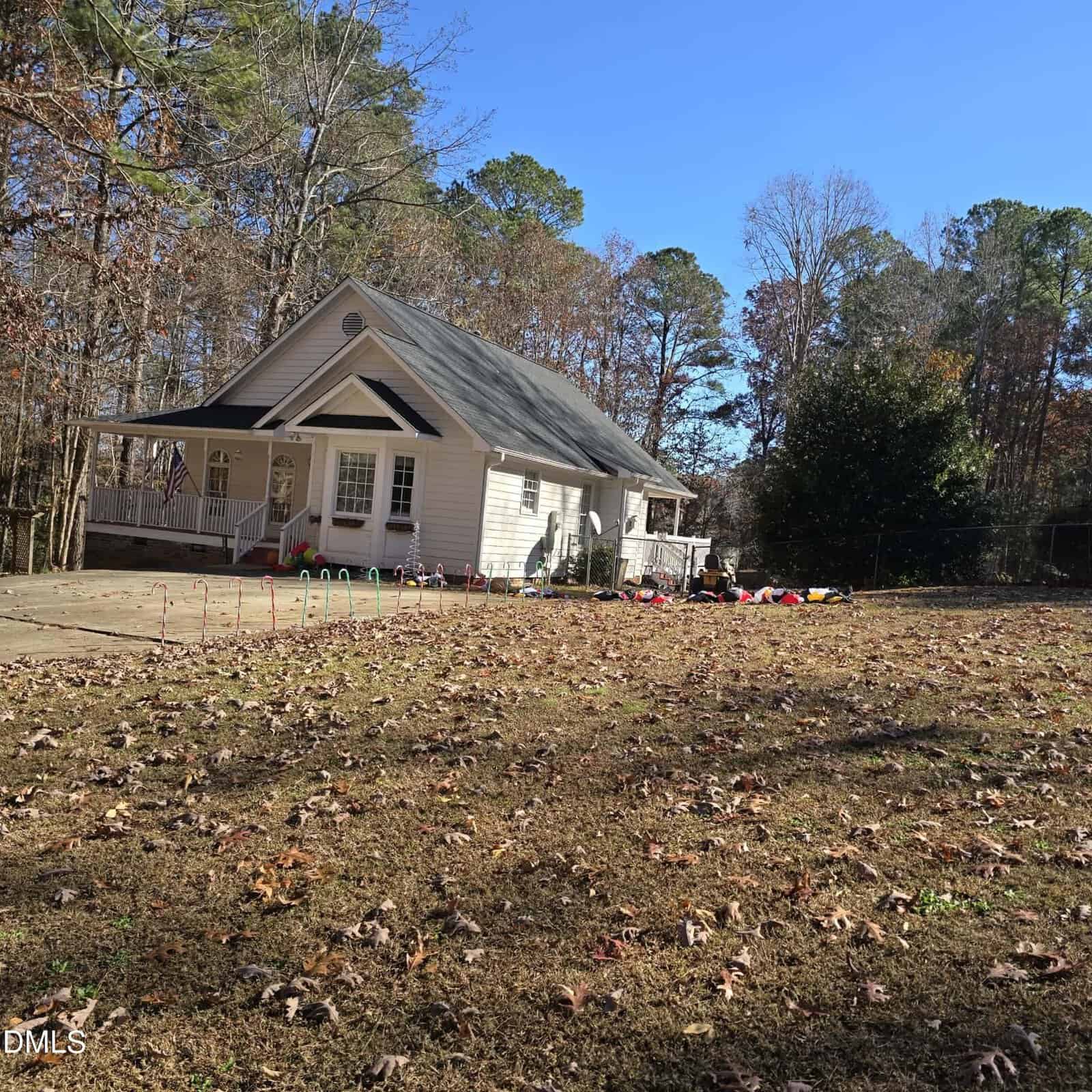 A white single-story house with a front porch sits on a large yard covered in dry leaves. Children's toys and play equipment are scattered near the house. There are trees with sparse leaves in the background under a clear blue sky.