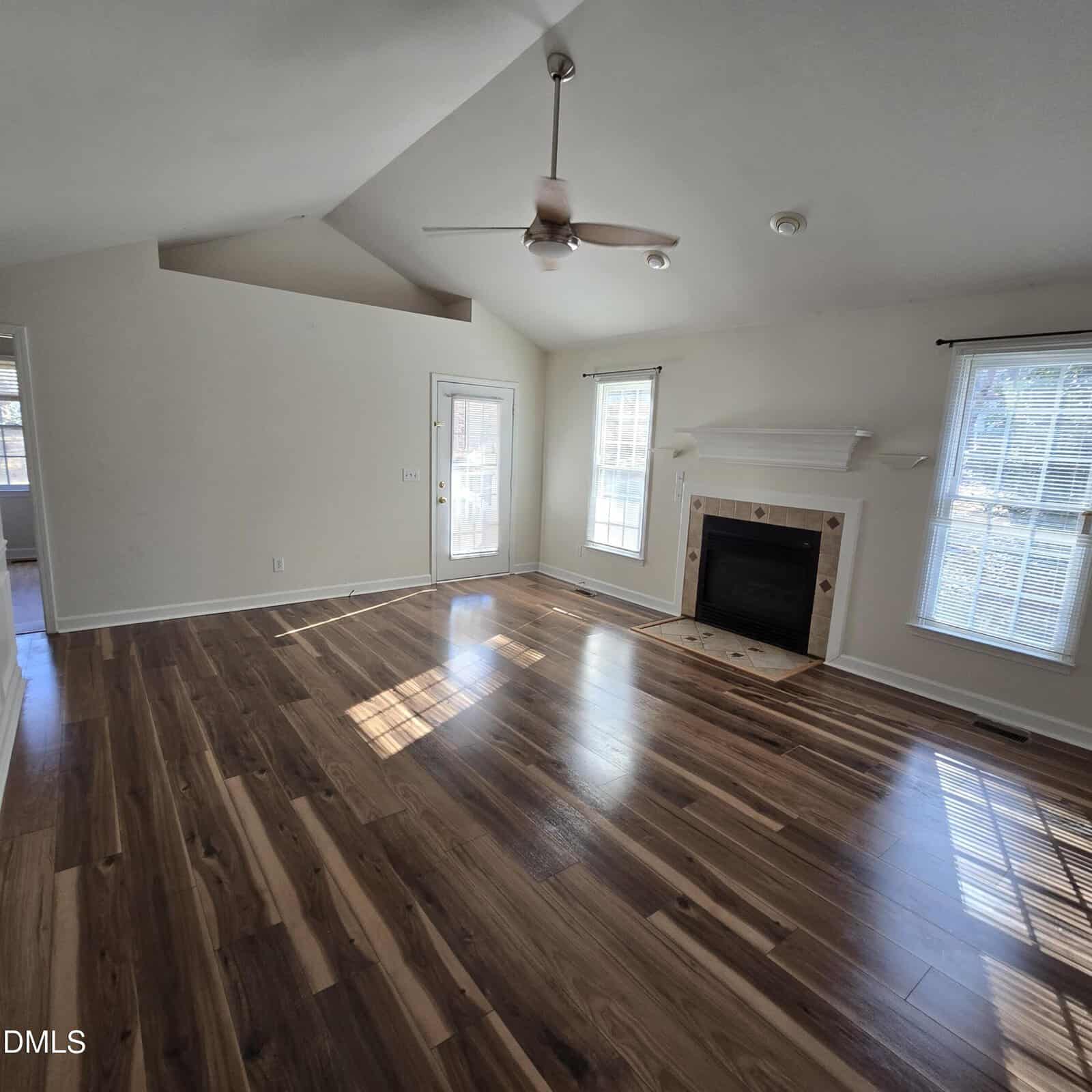 Bright living room with hardwood floors, a ceiling fan, white walls, a fireplace with a mantel, and two large windows letting in natural light. A glass door leads outside. Room is empty and unfurnished.