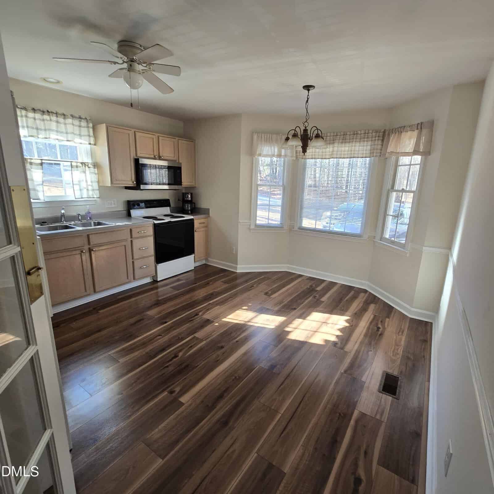A kitchen with light wood cabinets, a white stove, and a black dishwasher. There is a bay window area with a hanging light fixture and wood flooring throughout, with sunlight streaming in from the windows.